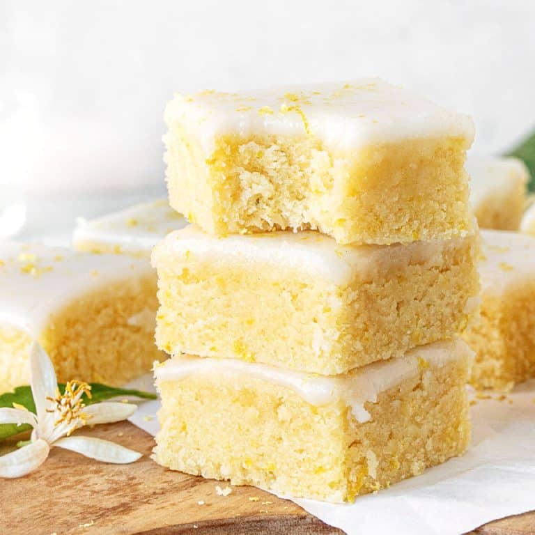 Three stacked glazed lemon brownies (or blondies) on a wooden board. Top one is bitten. White background.