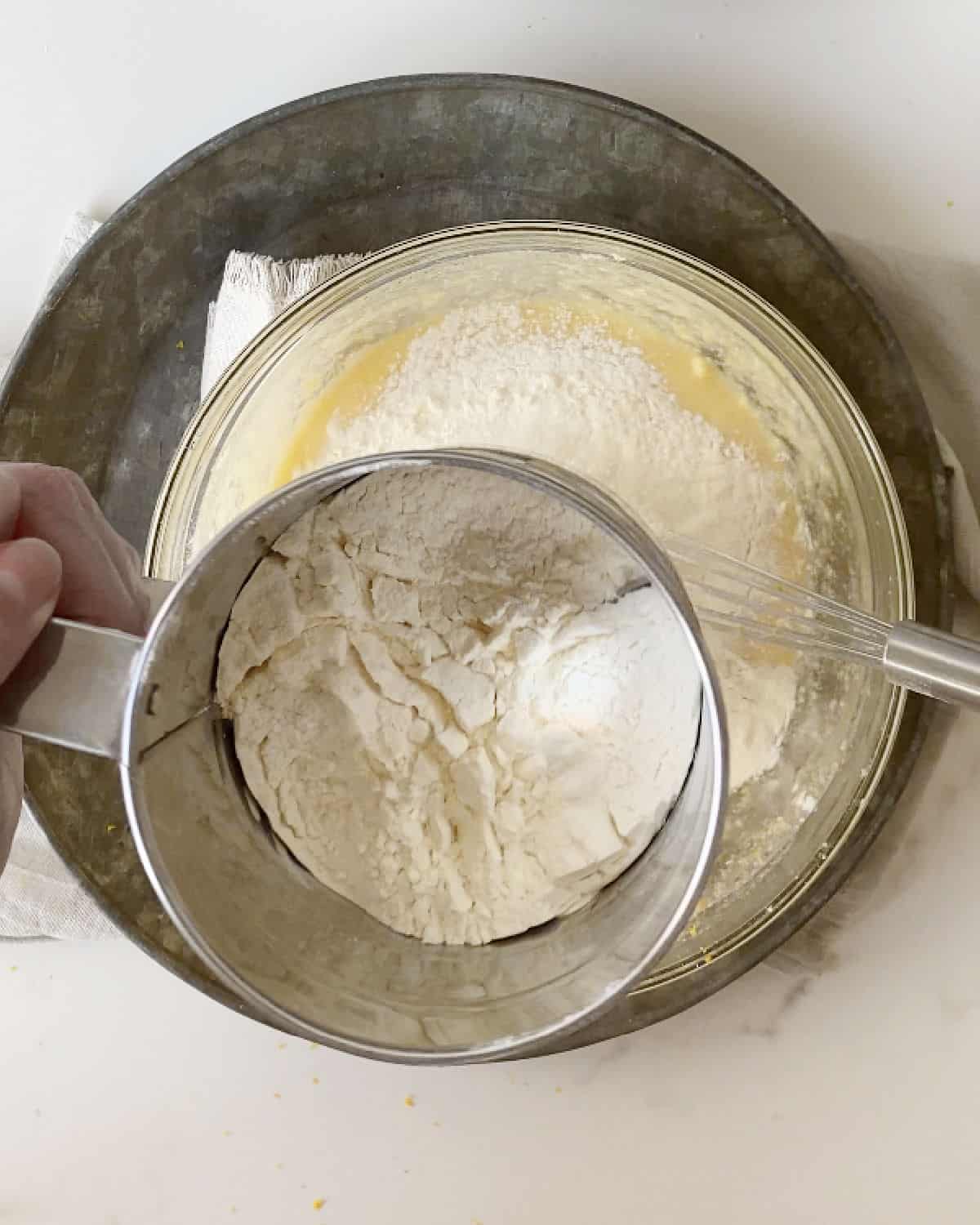 Flour being sifted over lemon batter in a glass bowl on a metal tray. White marble surface.