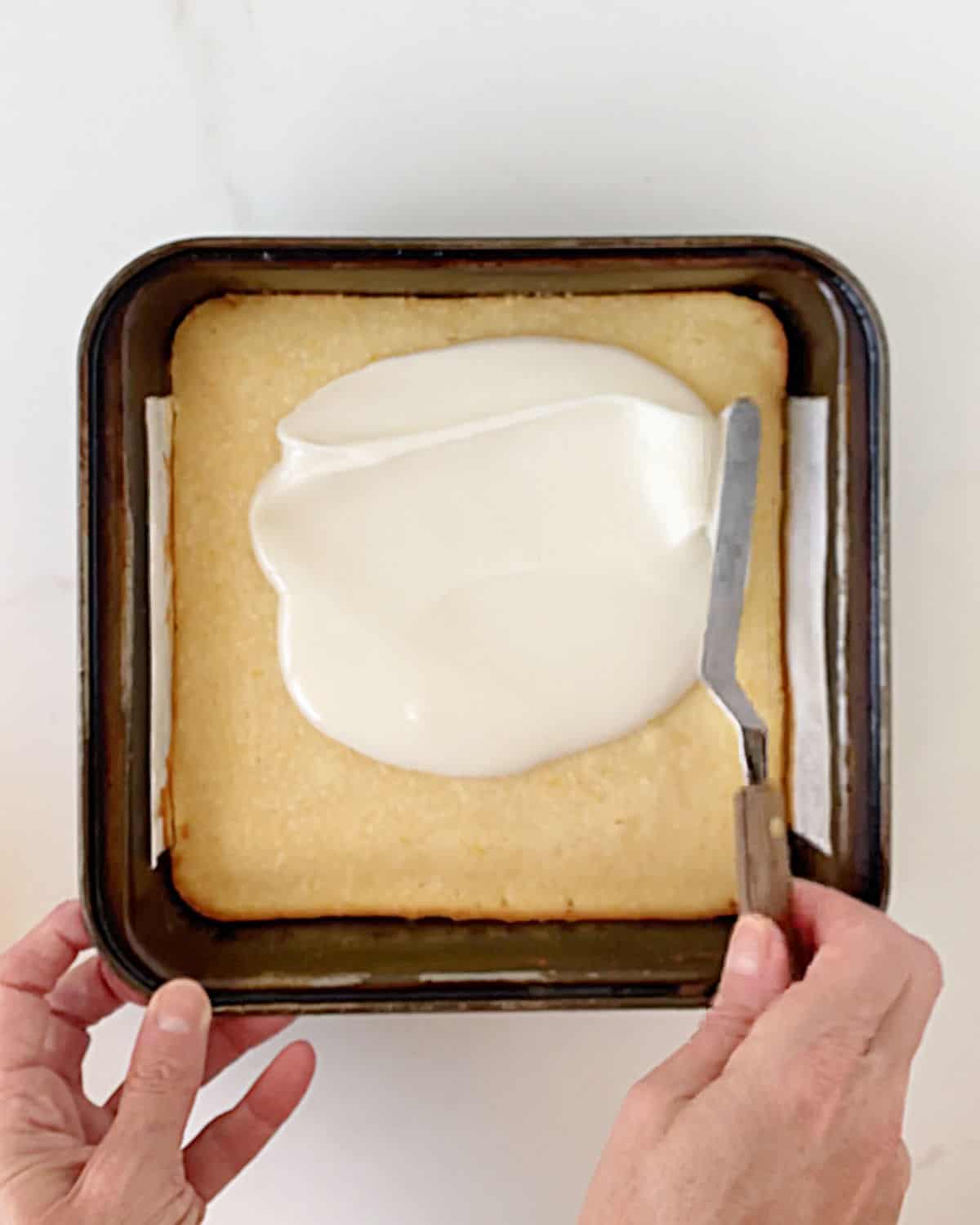 Glazing a square metal pan with lemon brownies on a white marble surface. 