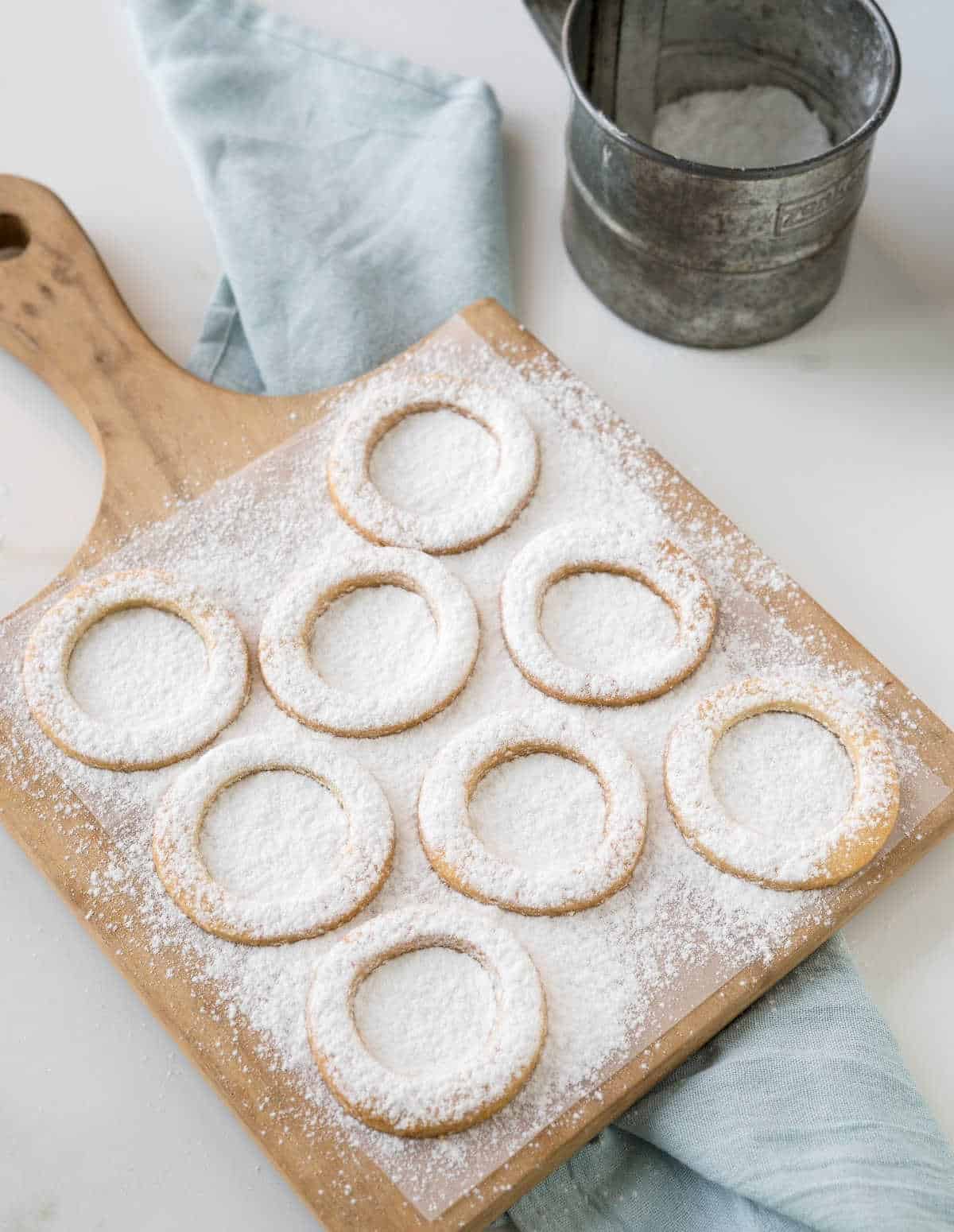 Wooden board with cookie rings covered in powdered sugar. A sifter and light blue cloth.