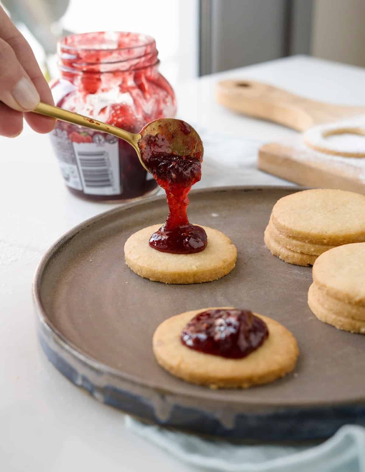 Placing a teaspoon of raspberry jam on cookie rounds on a brown plate. A jam jar. White surface.
