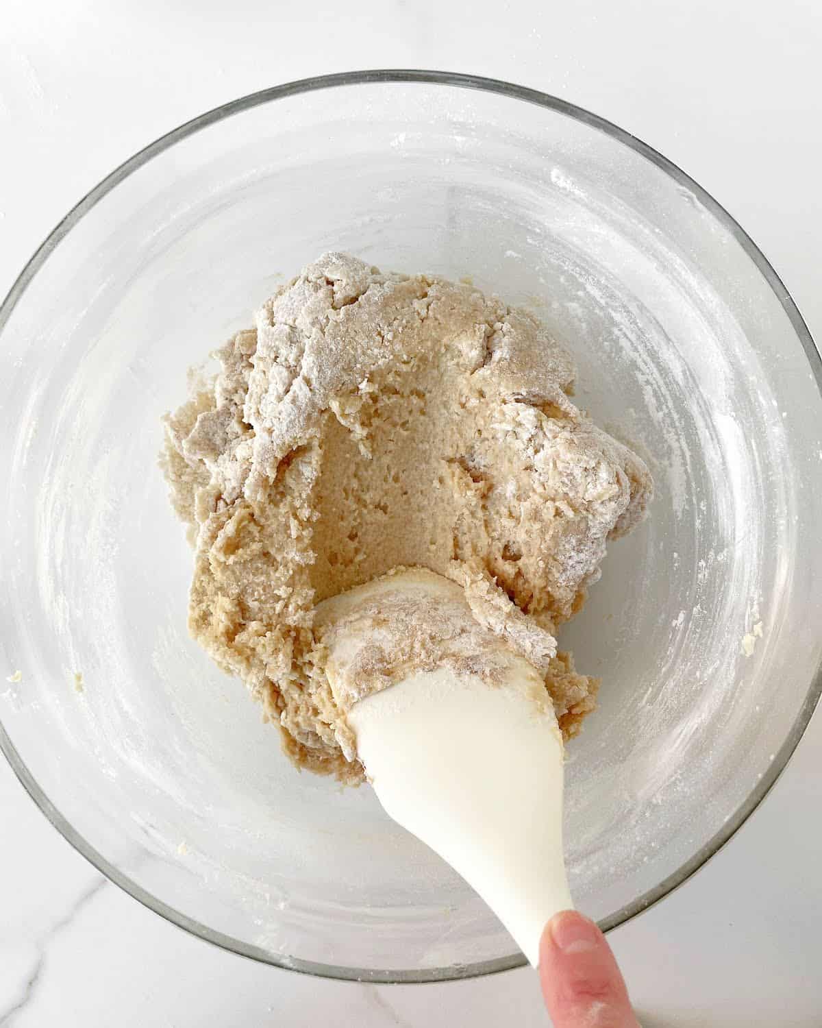 Flour being incorporated into cookie dough mixture with a white spatula in a glass bowl on white marble.
