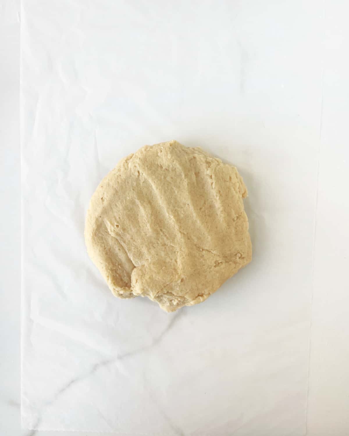 Disc of almond cookie dough on a piece of plastic on a white marble surface.