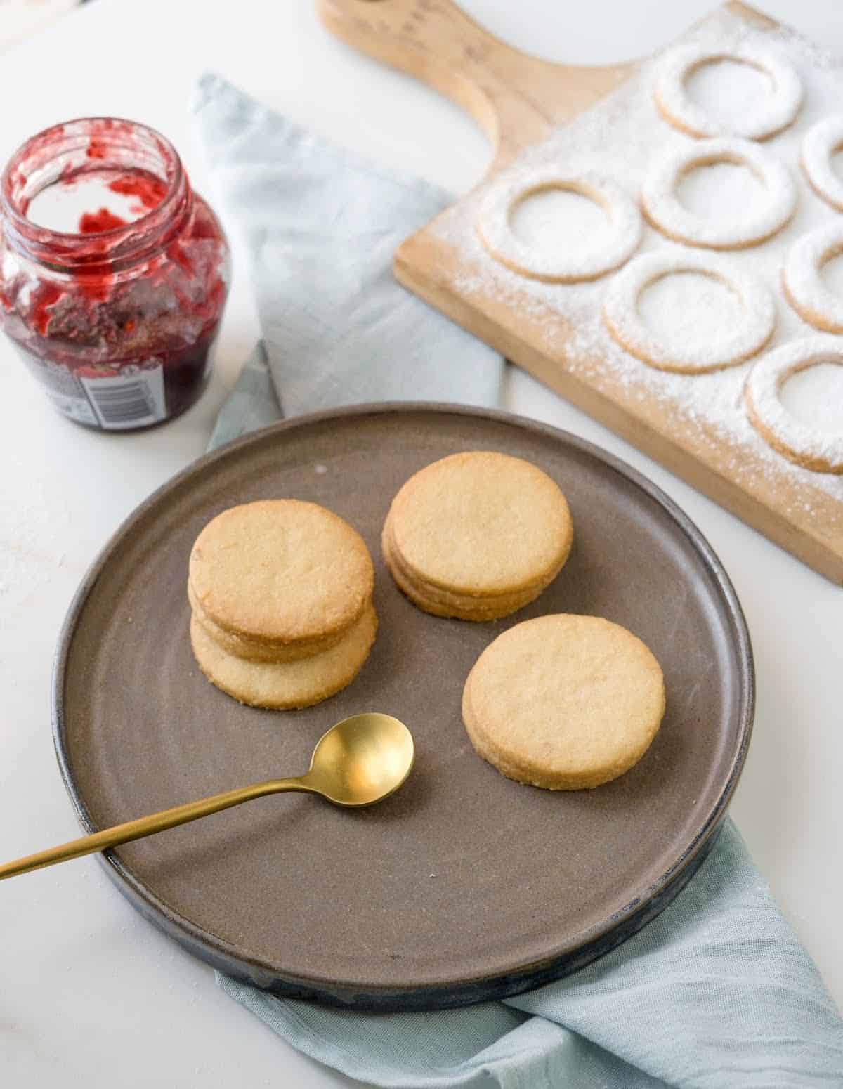 Brown plate with almond cookies on a white surface with a blue cloth. Jar of jam and wooden board with cookie rings.