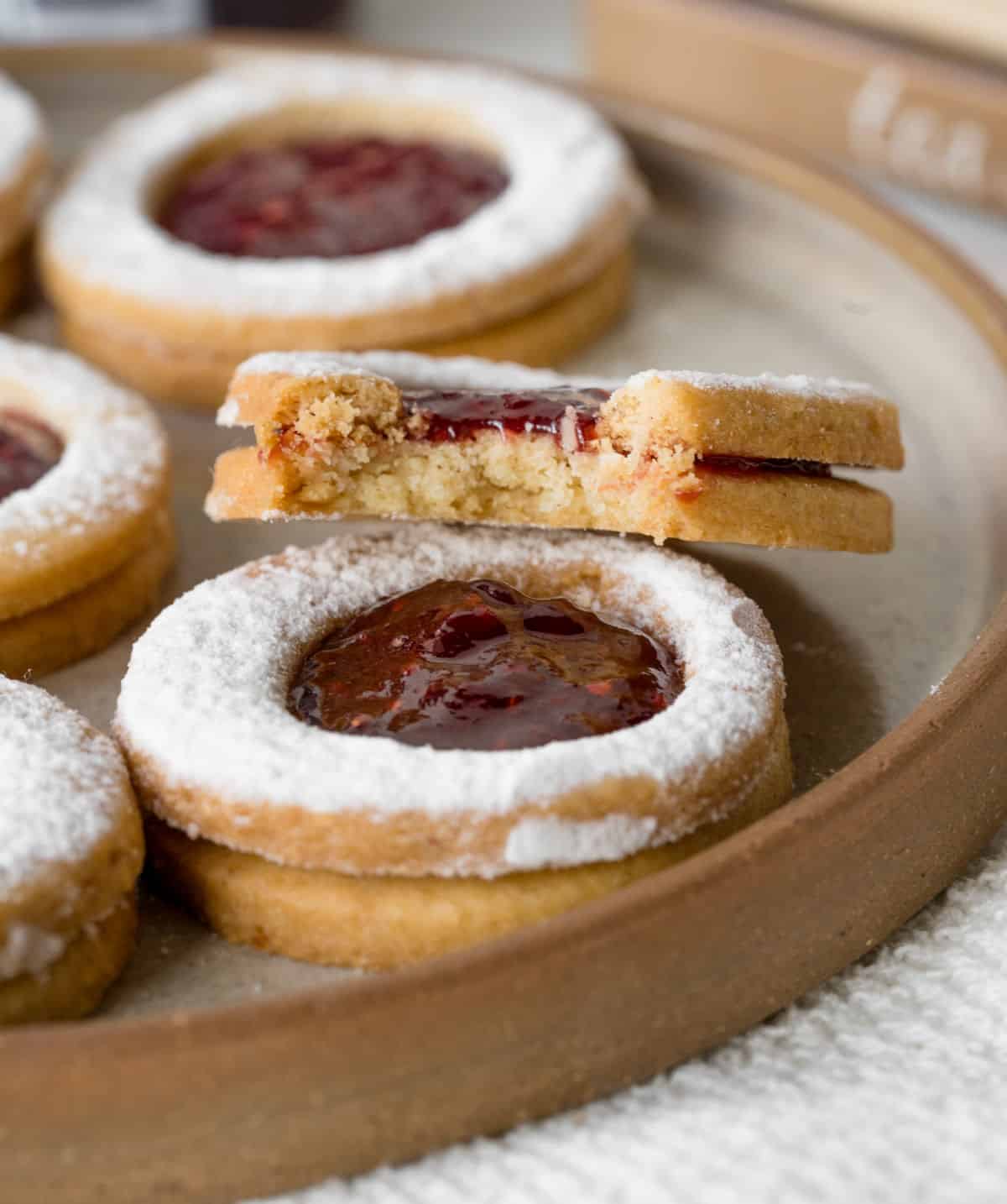 Eaten raspberry linzer cookie leaning on other whole cookies on a brown plate. 