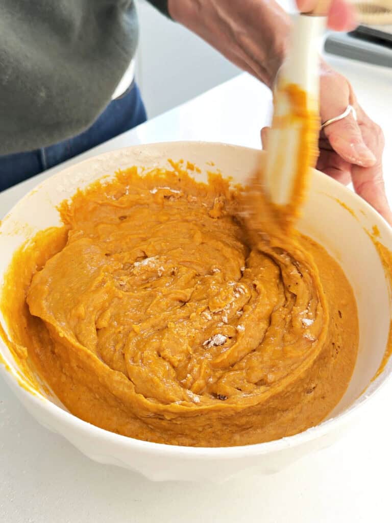 Stirring pumpkin maple muffin batter in a white bowl with a white spatula.