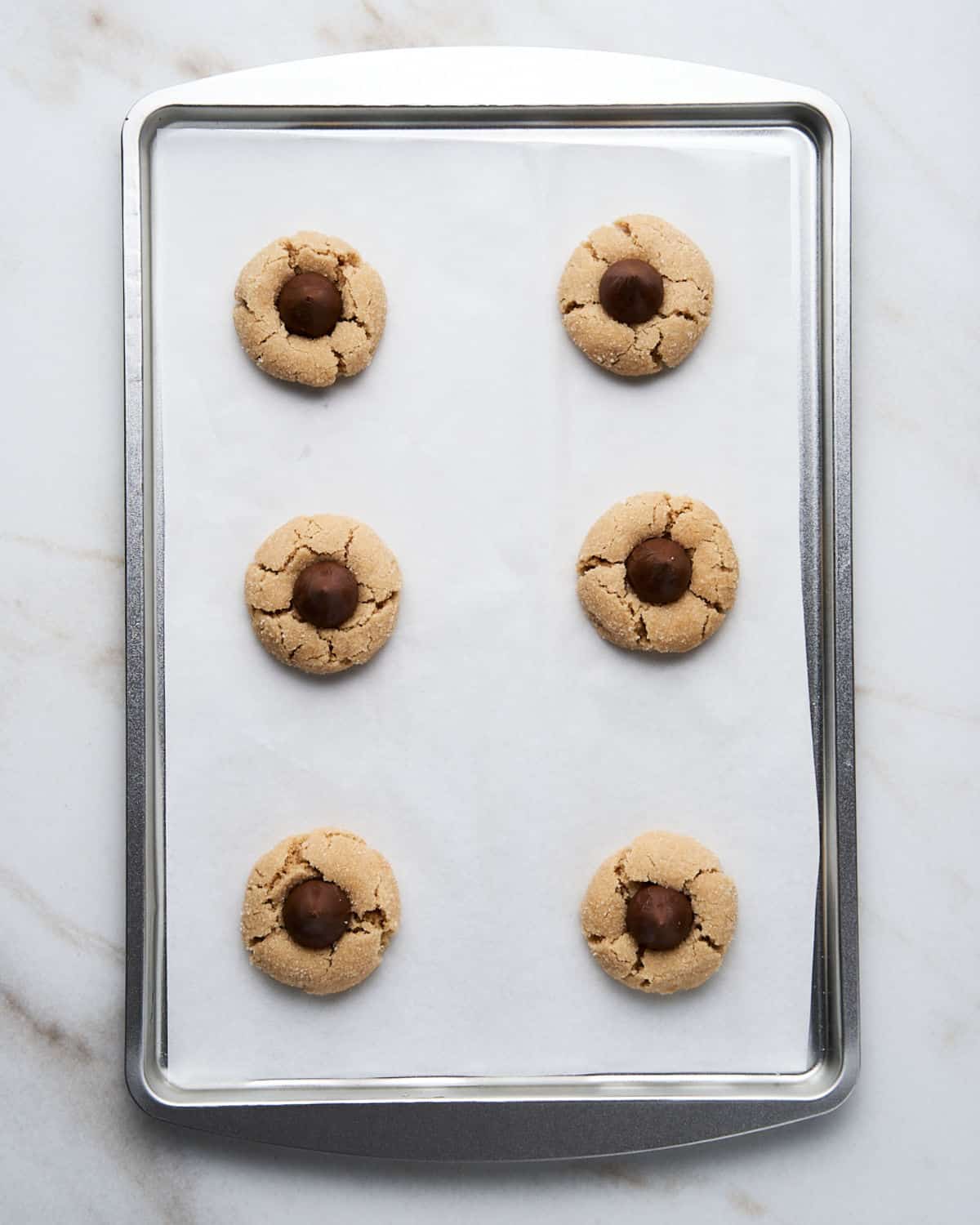 Baked peanut butter blossom cookies on white parchment paper on a metal cookie sheet placed on a white marble counter.