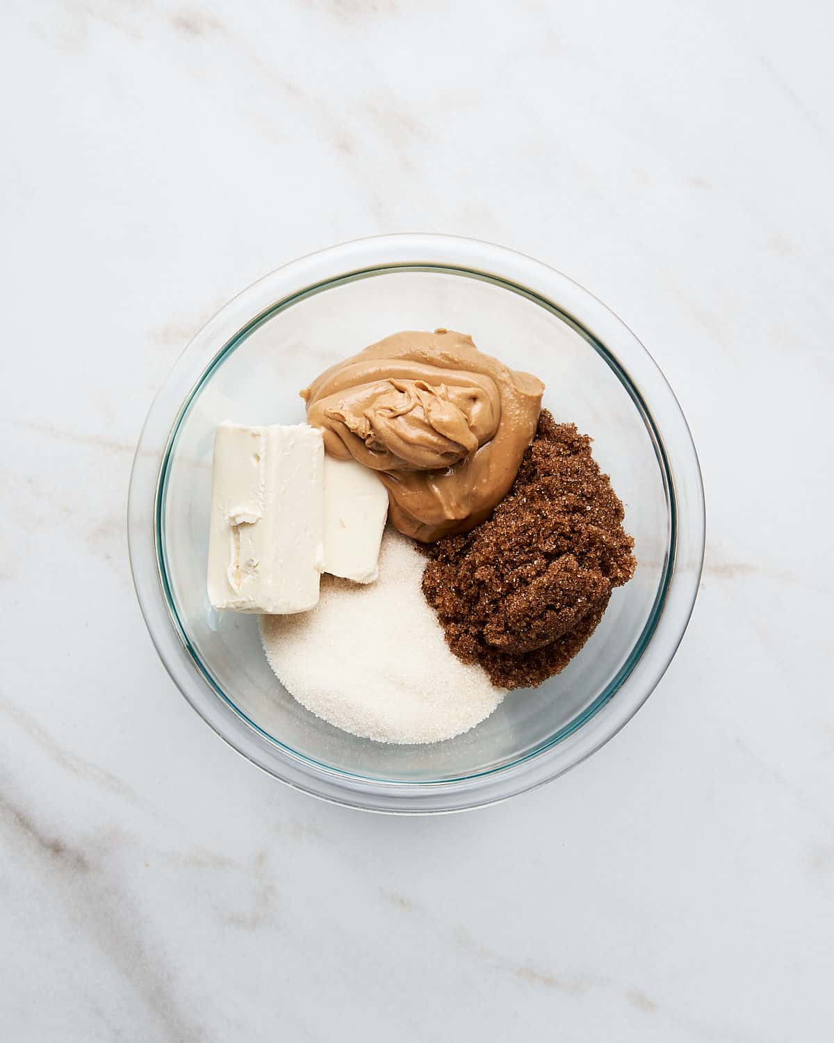 Brown sugar, butter, sugar, and peanut butter in a glass bowl on a white marble surface.