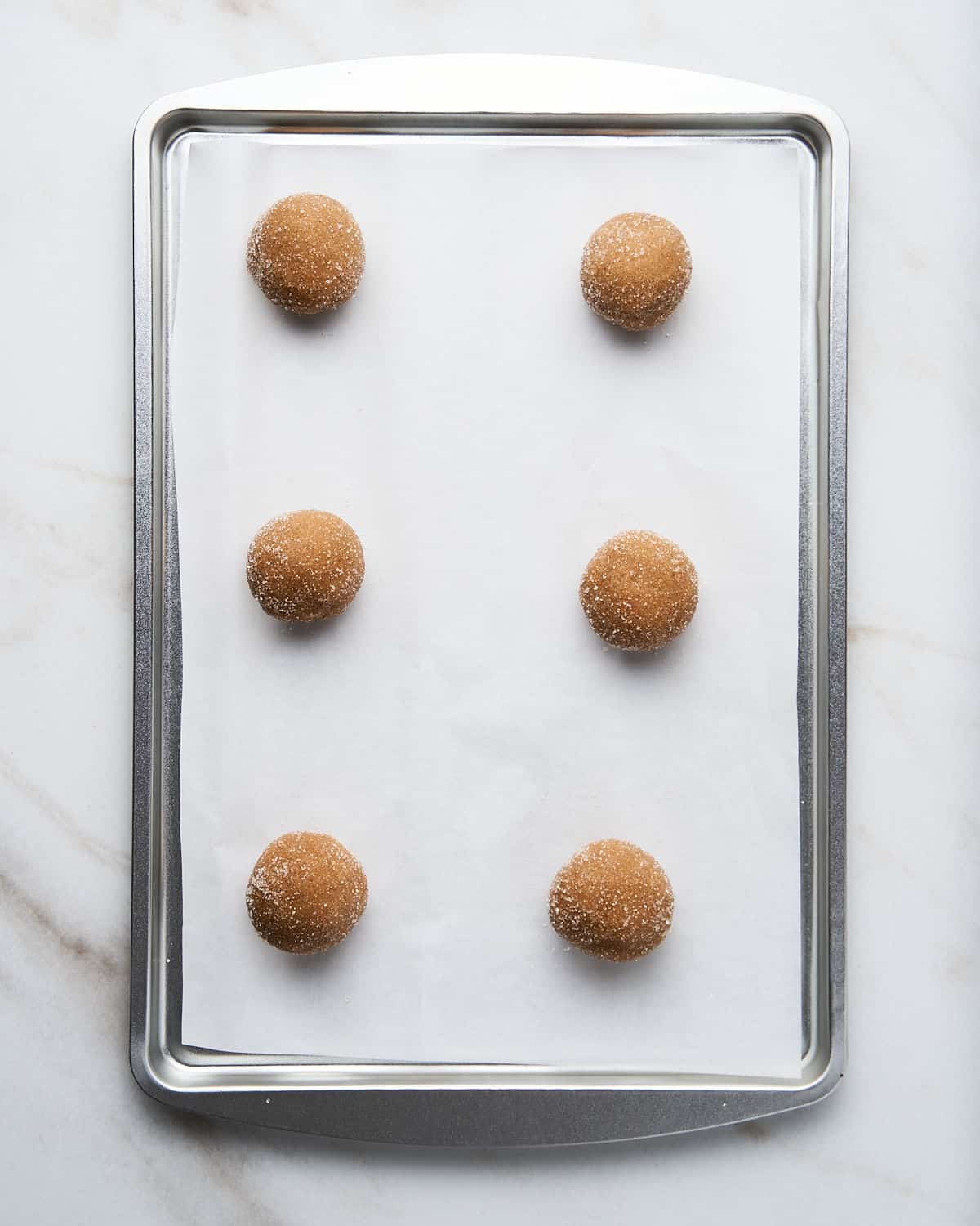 Six peanut butter cookie dough balls on white parchment paper on a cookie sheet. White marble surface.