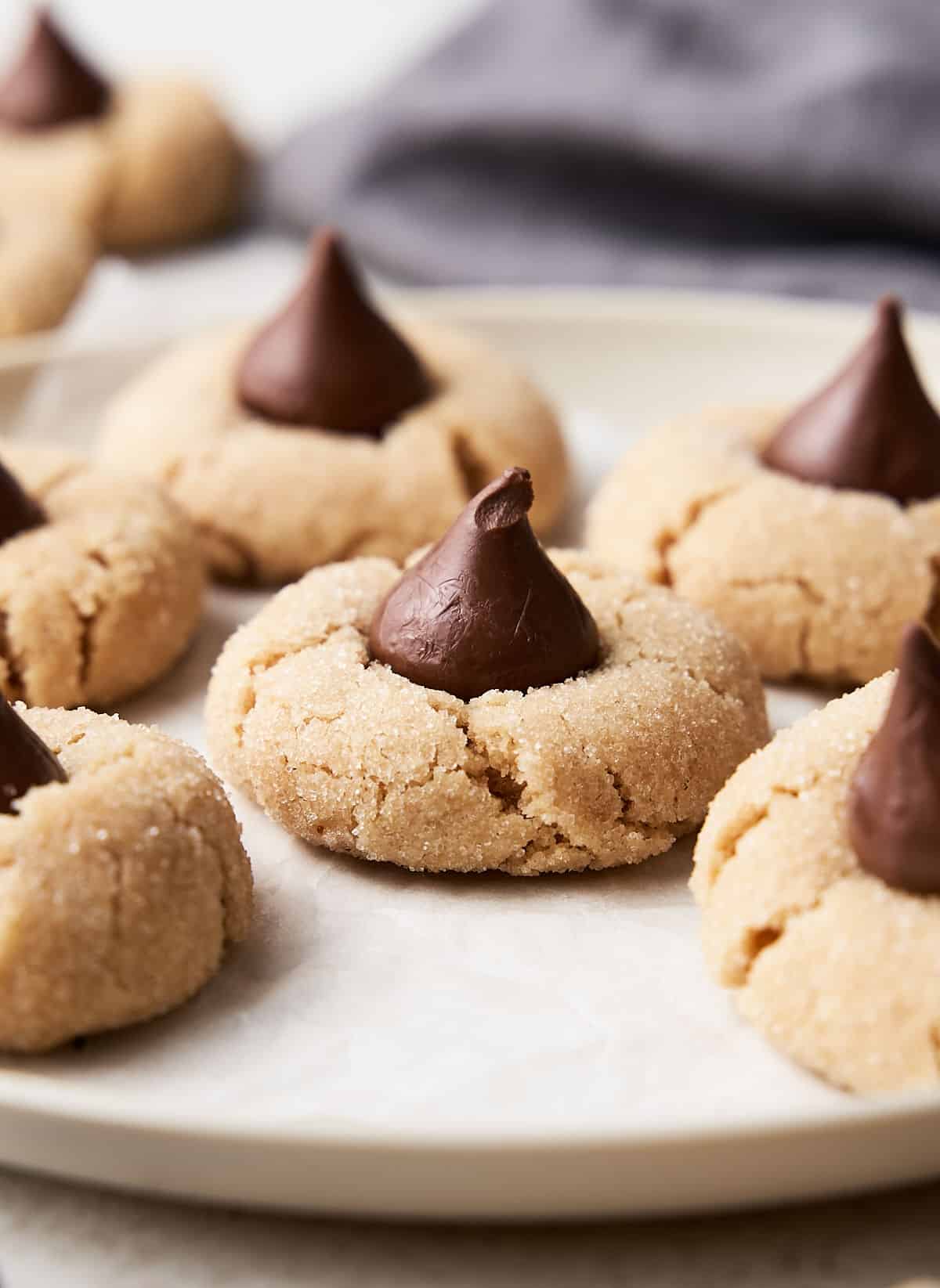Cream colored plate with several peanut butter cookies with chocolate kisses. Grey cloth in the backbground.