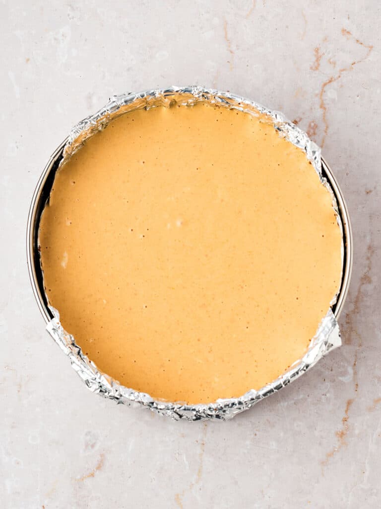 Unbaked pumpkin cheesecake batter in a foil-lined round pan. White marbled surface.