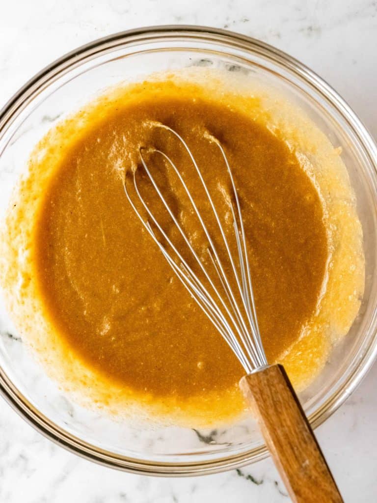 Mixed wet ingredients for pumpkin muffins batter in a glass bowl with a hand whisk inside. White marble surface.