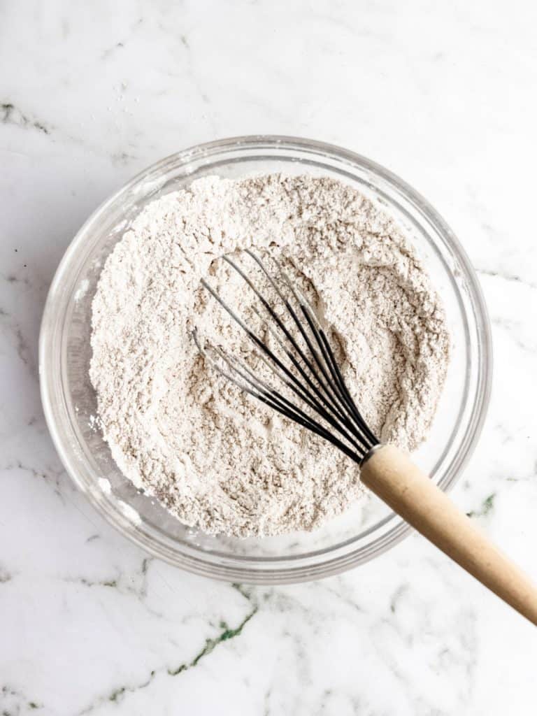 Dry ingredients for muffins in a glass bowl with a whisk on a white marble surface.