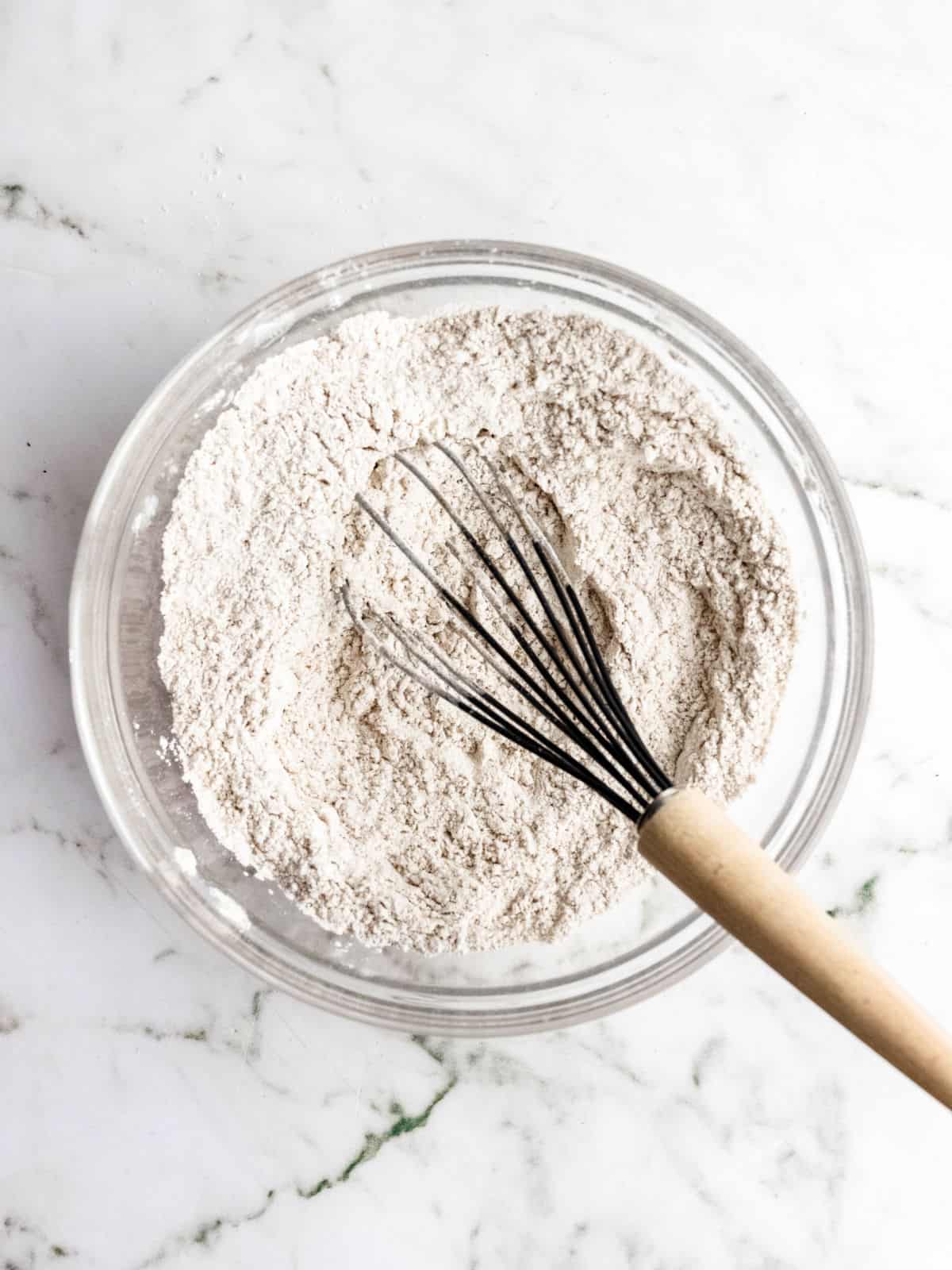 Dry ingredients for muffins in a glass bowl with a whisk on a white marble surface.