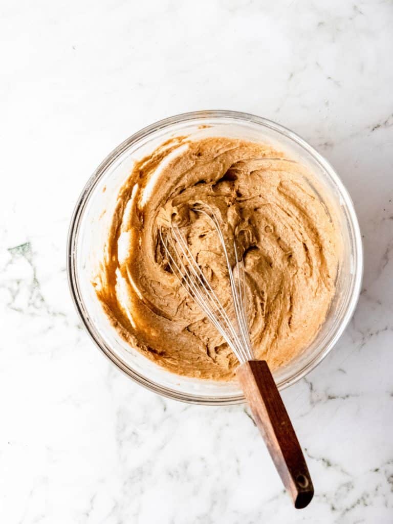 Pumpkin muffin batter in a glass bowl with a whisk on a white marble surface. View from the top.