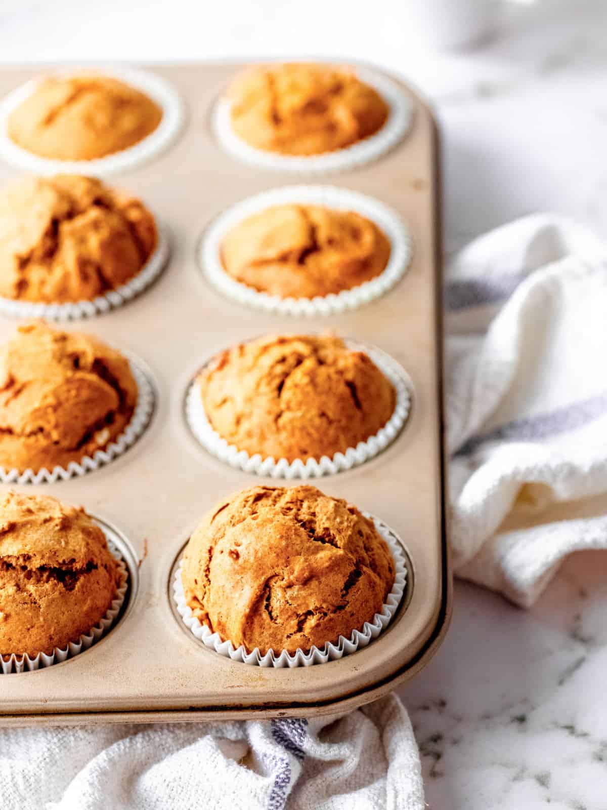 Baked pumpkin muffins in a muffin pan on a white kitchen cloth on a white marble surface. .