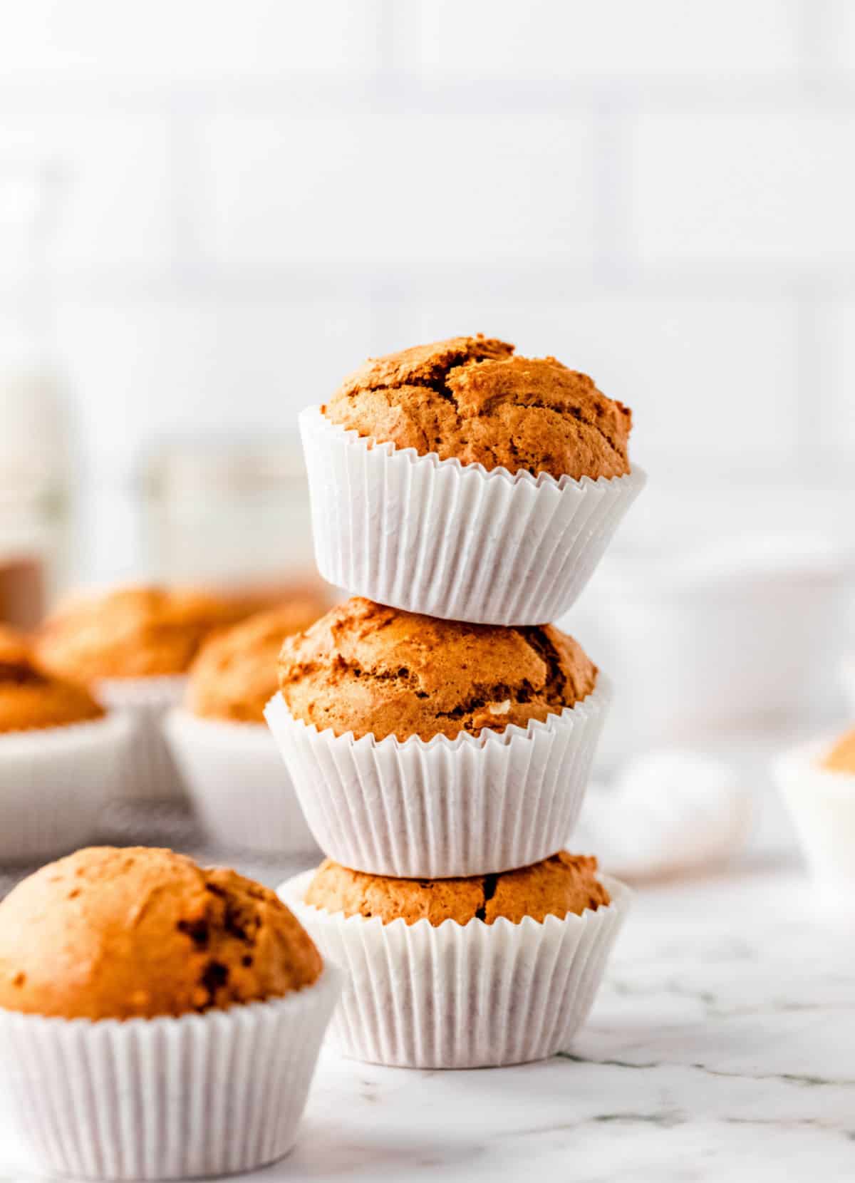 Stack of three pumpkin muffins on a white surface and background with more muffins around.
