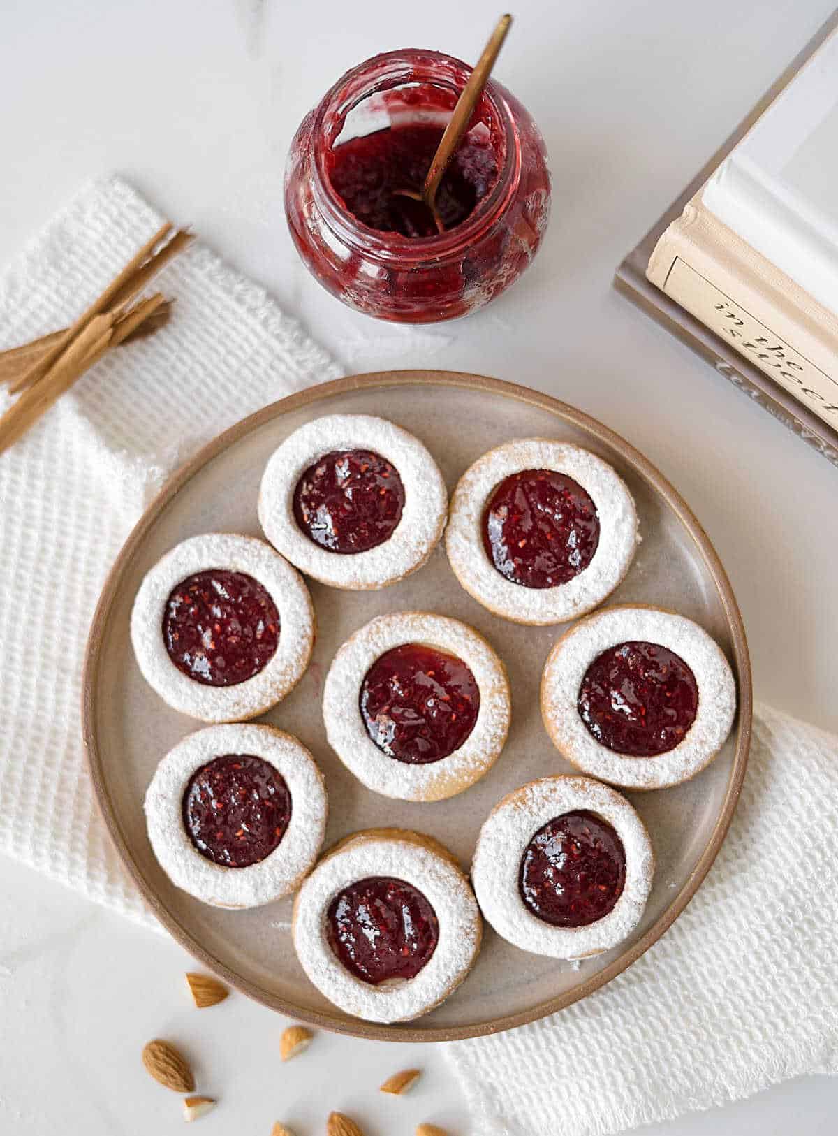 Several raspberry linzer cookies on a brown plate. White surface, white kitchen towel. A jam jar and cinnamon sticks.