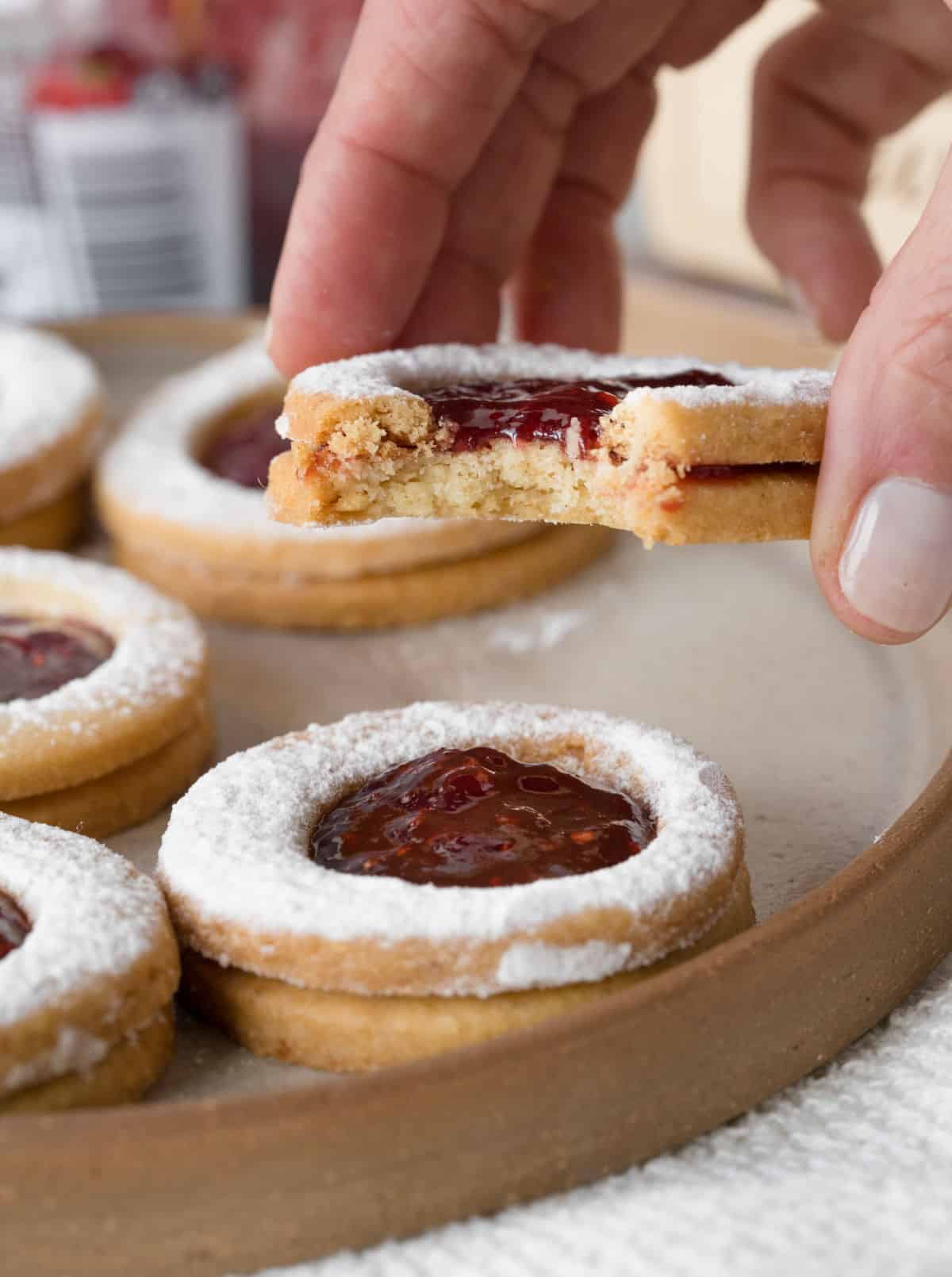 Hand holding bitter linzer cookie with several others whole on a beige tray below. 