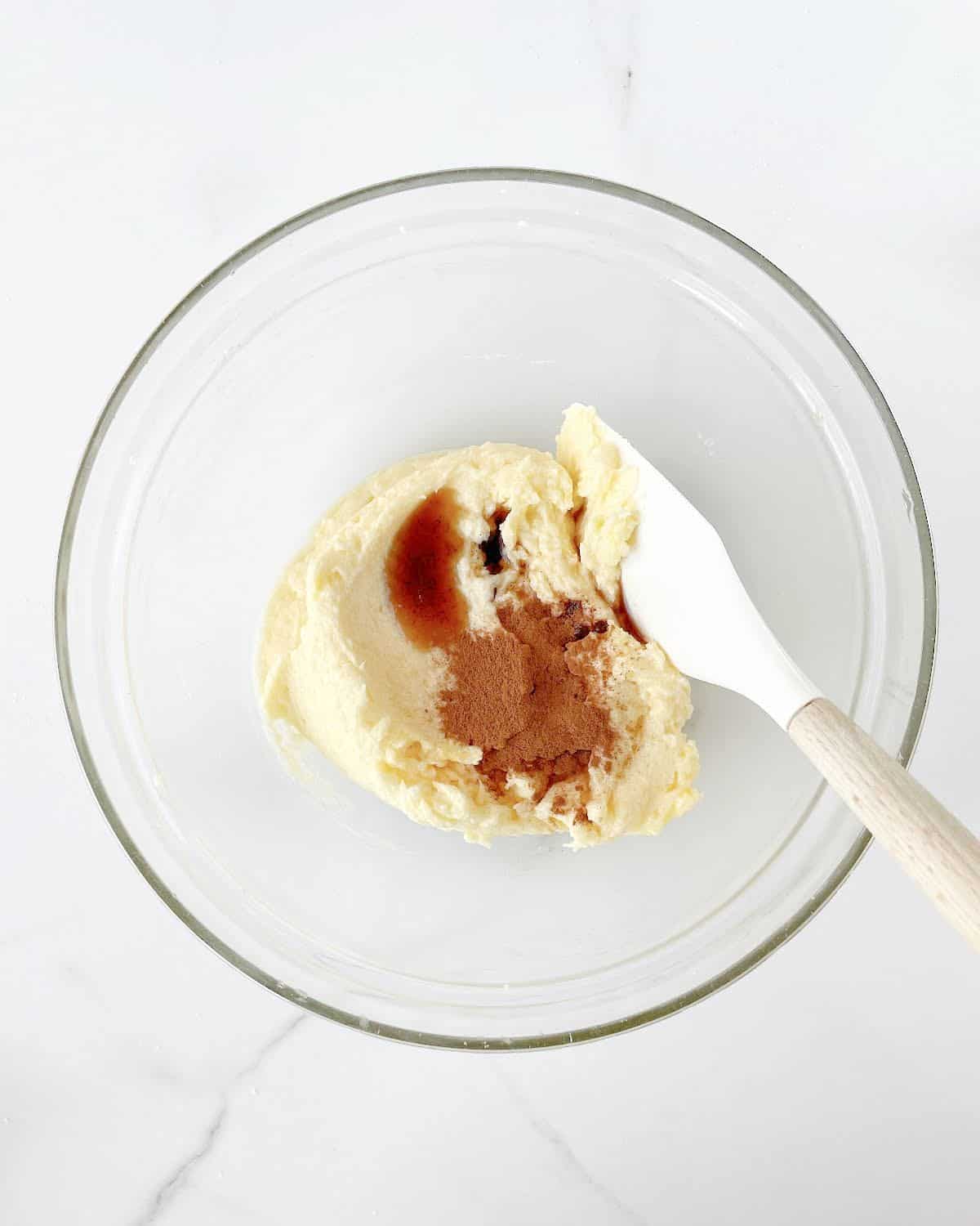 Butter mixture with vanilla and ground cinnamon in a glass bowl with a white spatula. White marble surface. 