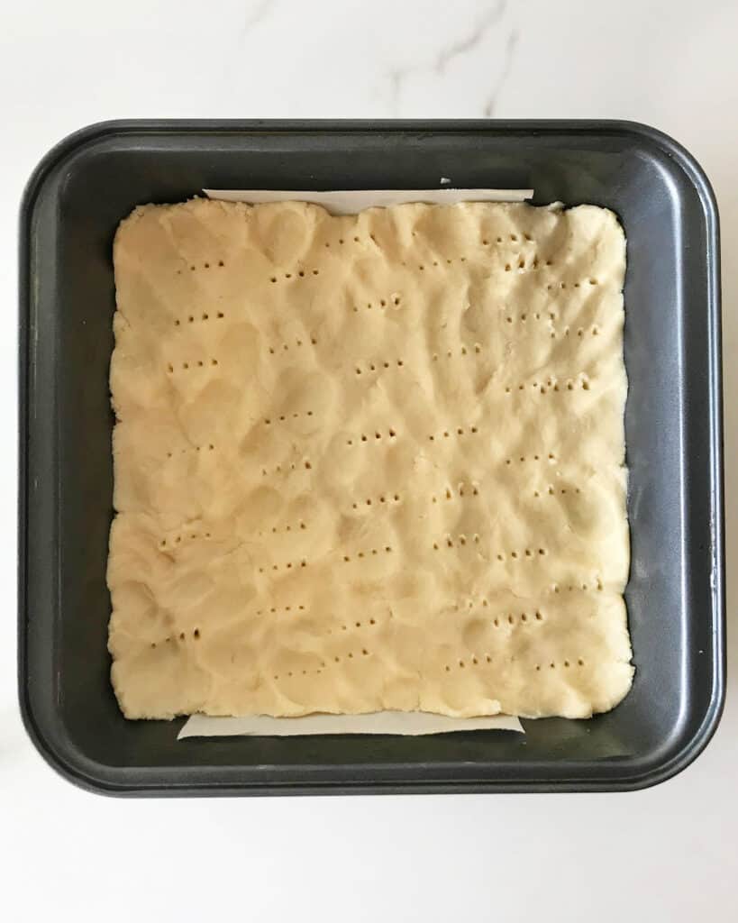 Metal square pan with shortbread crust before baking. White background.