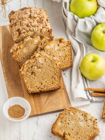 View from above of wooden board on a white surface with apple bread slices, a kitchen cloth, green apples, cinnamon sugar in a bowl.