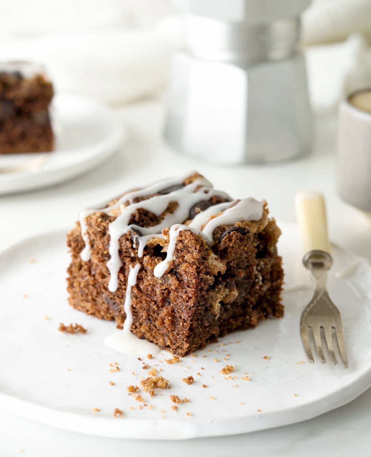 Square of glazed chocolate coffee cake on a white plate with fork. White background with coffee pot.