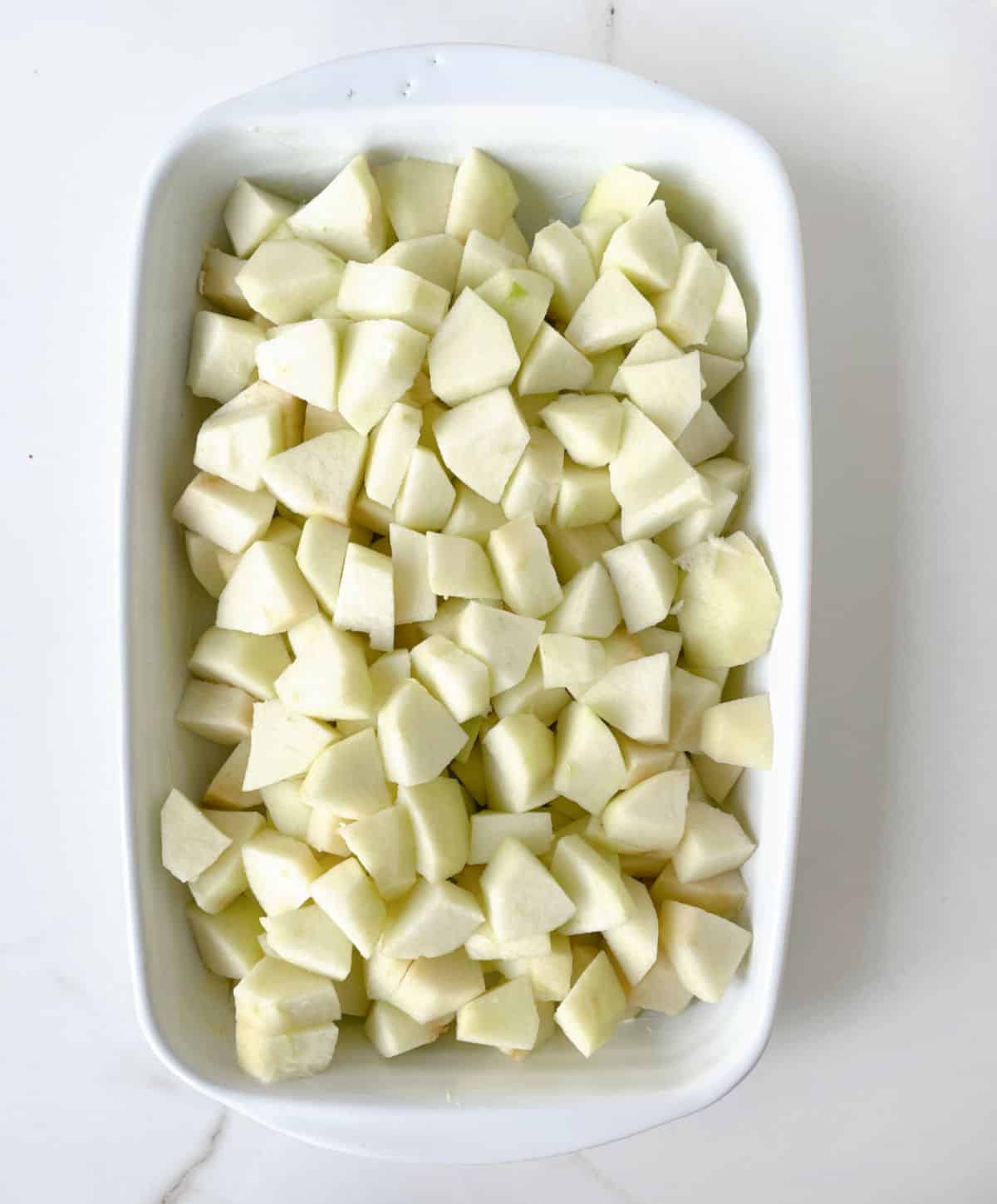 Pieces of apple in a white baking dish on a white marble surface. 