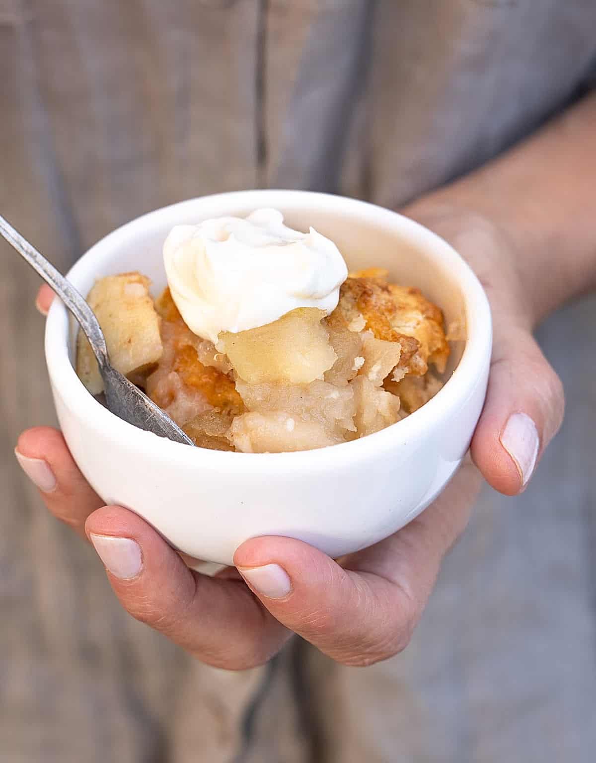 Serving of apple cobbler with dollop of cream in a white bowl being held. Grey shirt as background. 