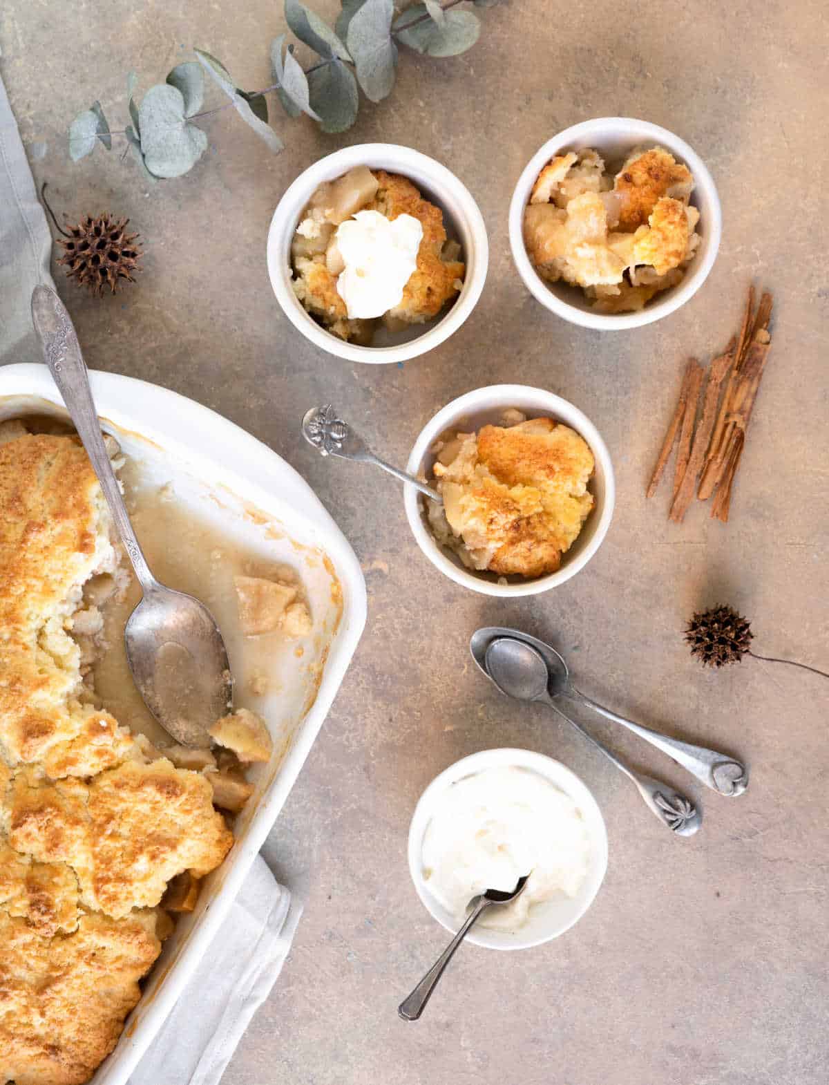 Three white bowls and baking pan with apple cobbler. Spoons, cinnamon sticks, brown surface. 