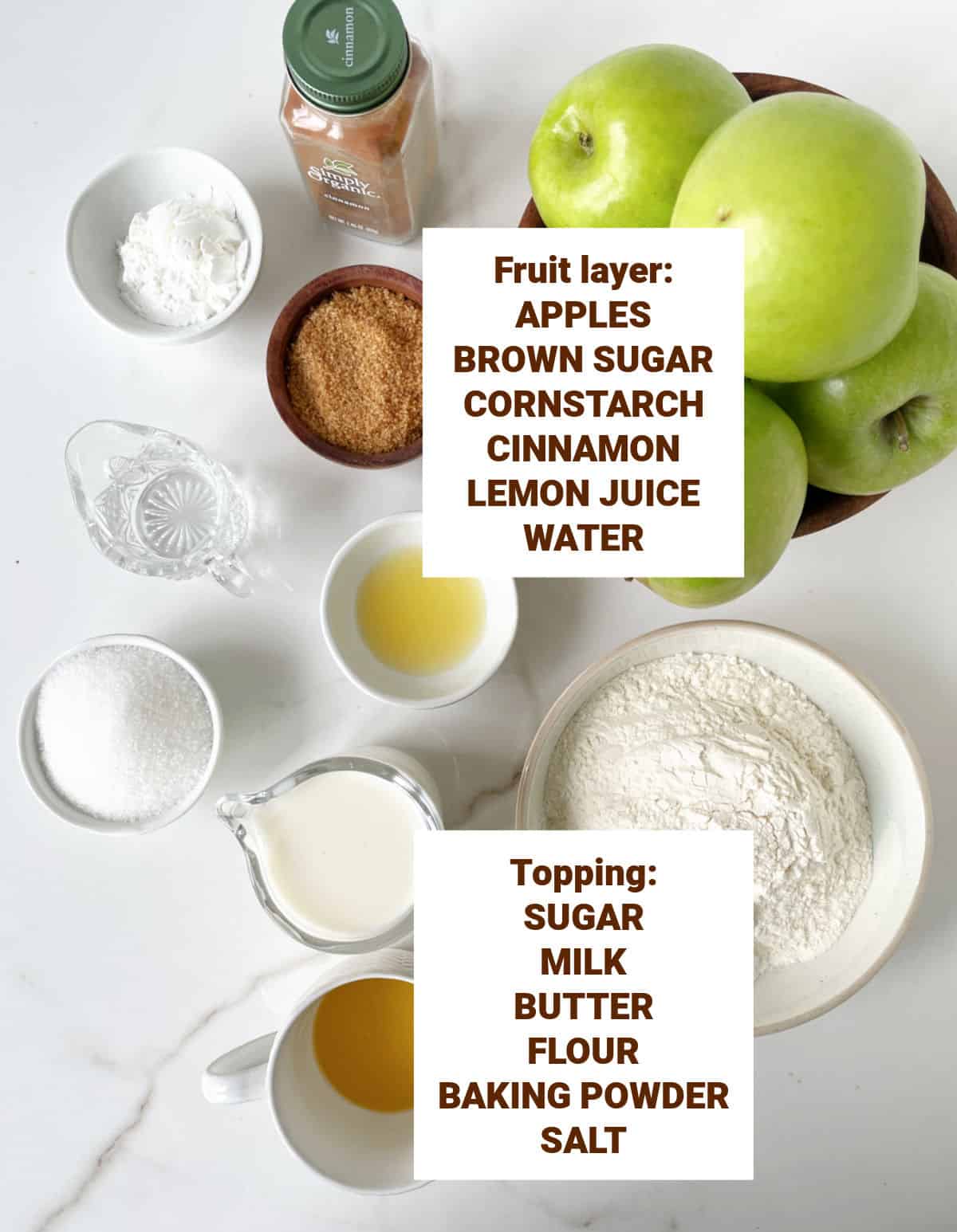White marble surface with bowls containing ingredients for apple cobbler including sugars, flour, butter, cinnamon, cornstarch, lemon juice, milk.