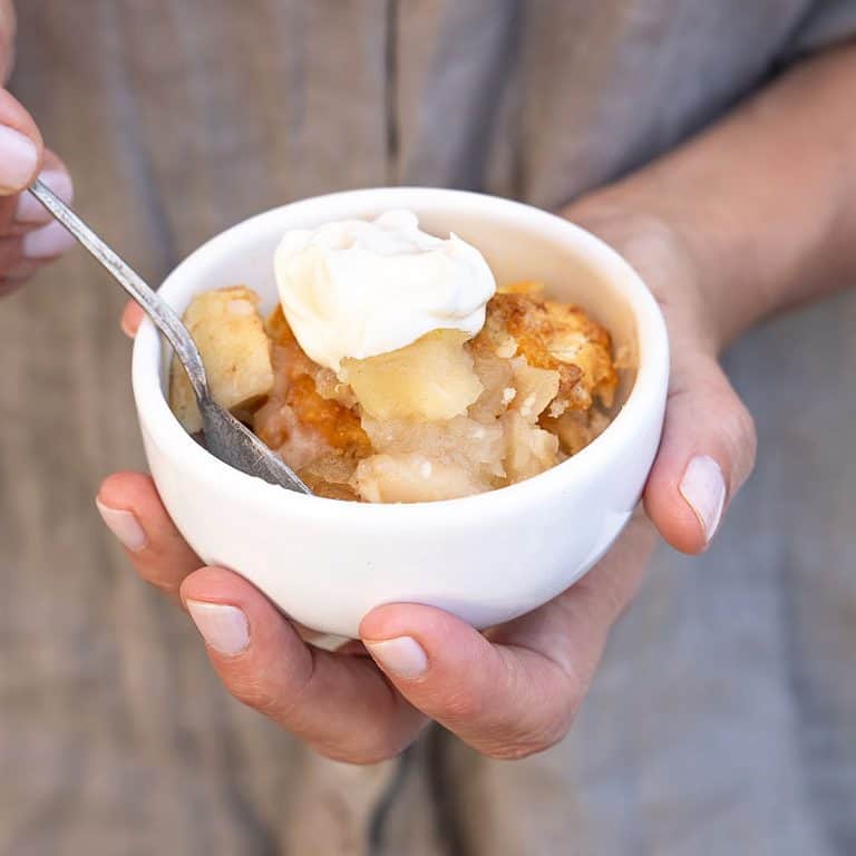 Hand holding white bowl with apple cobbler with whipped cream. Grey background.