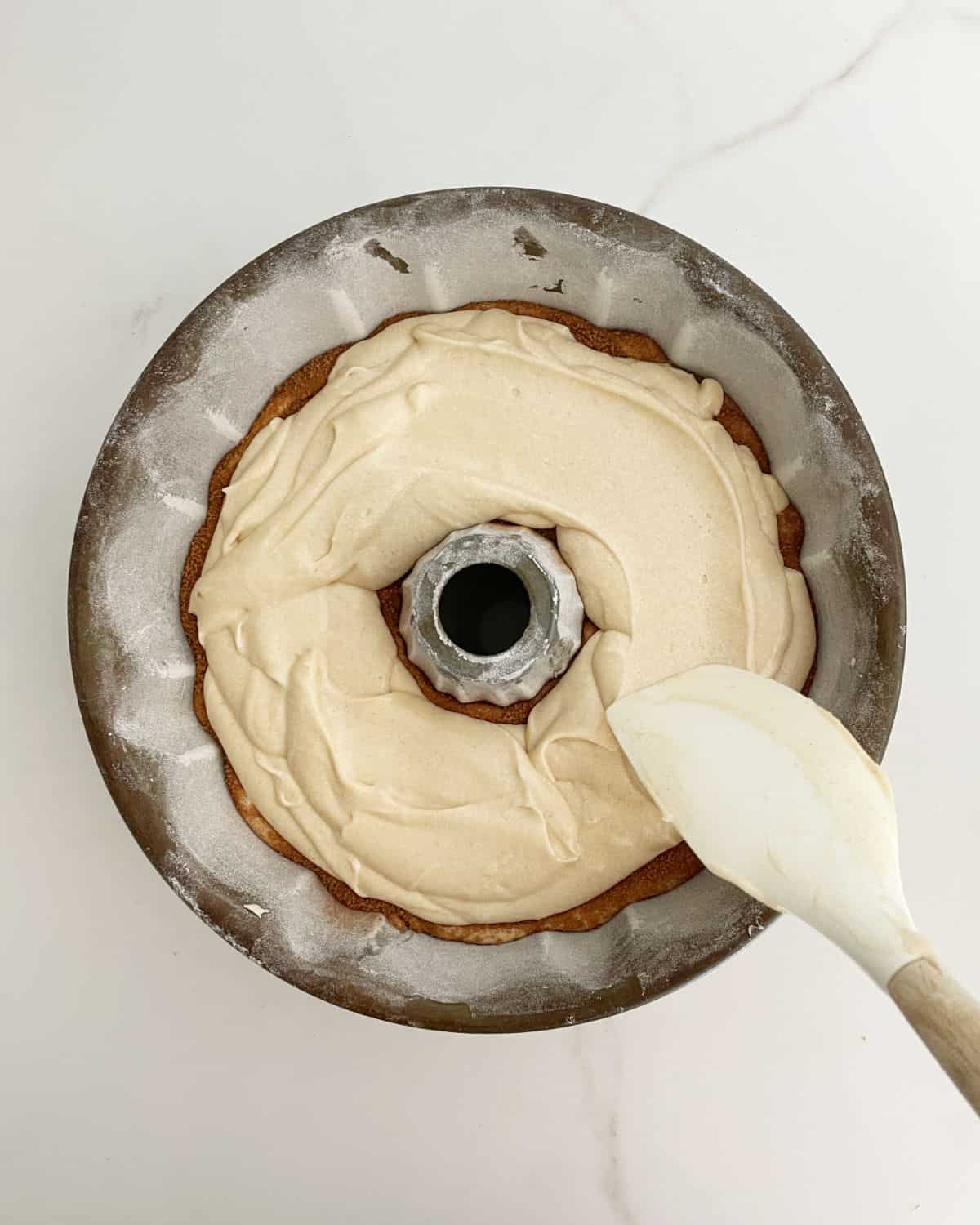 Top view of bundt cake pan with cinnamon cake batter being smoothed with a white spatula. White marble surface.