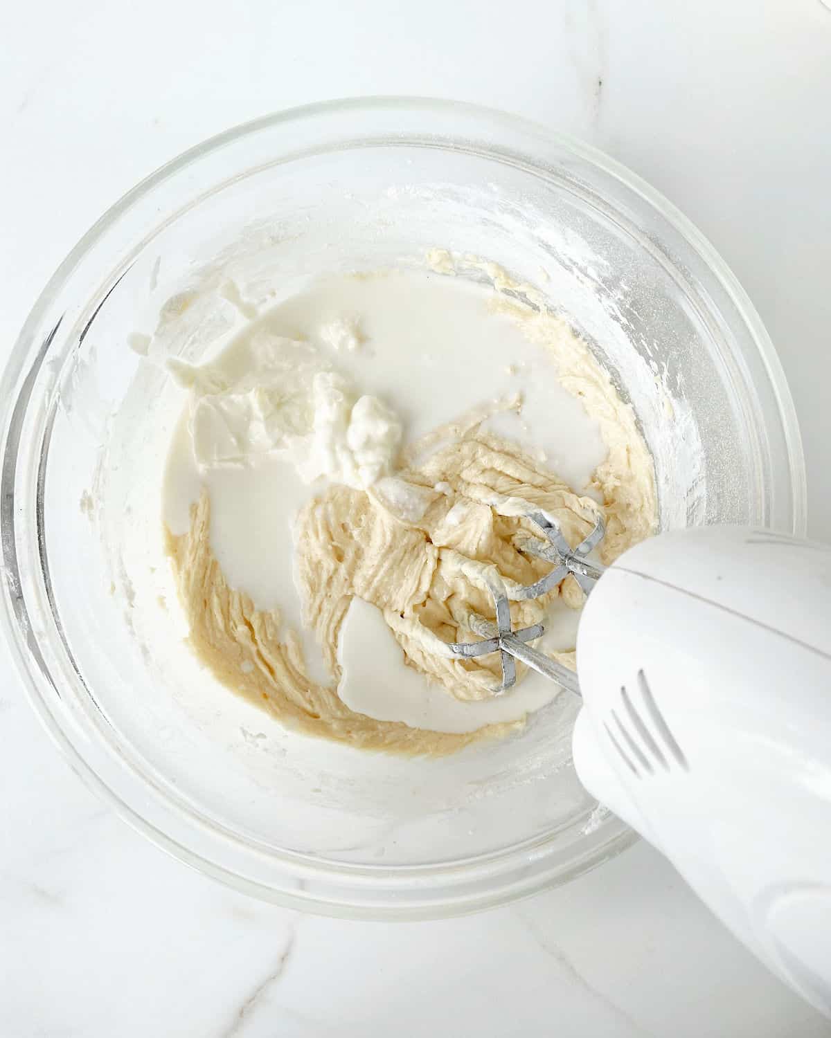 Milk and sour cream added to cake batter in a glass bowl with an electric mixer. White marble surface.