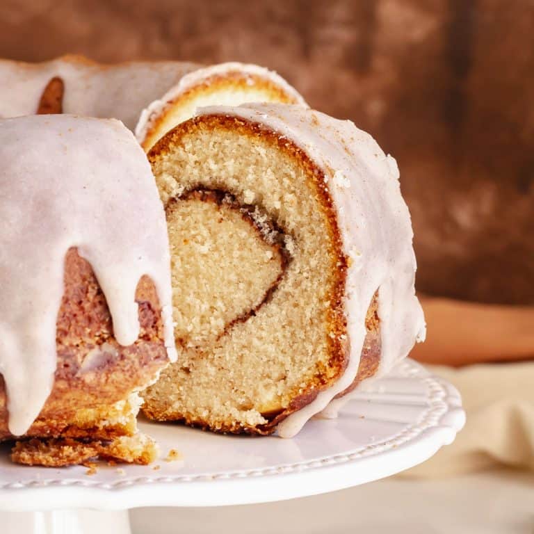 Slice and whole cake of glazed cinnamon swirl bundt cake on a white cake stand. Brown background.