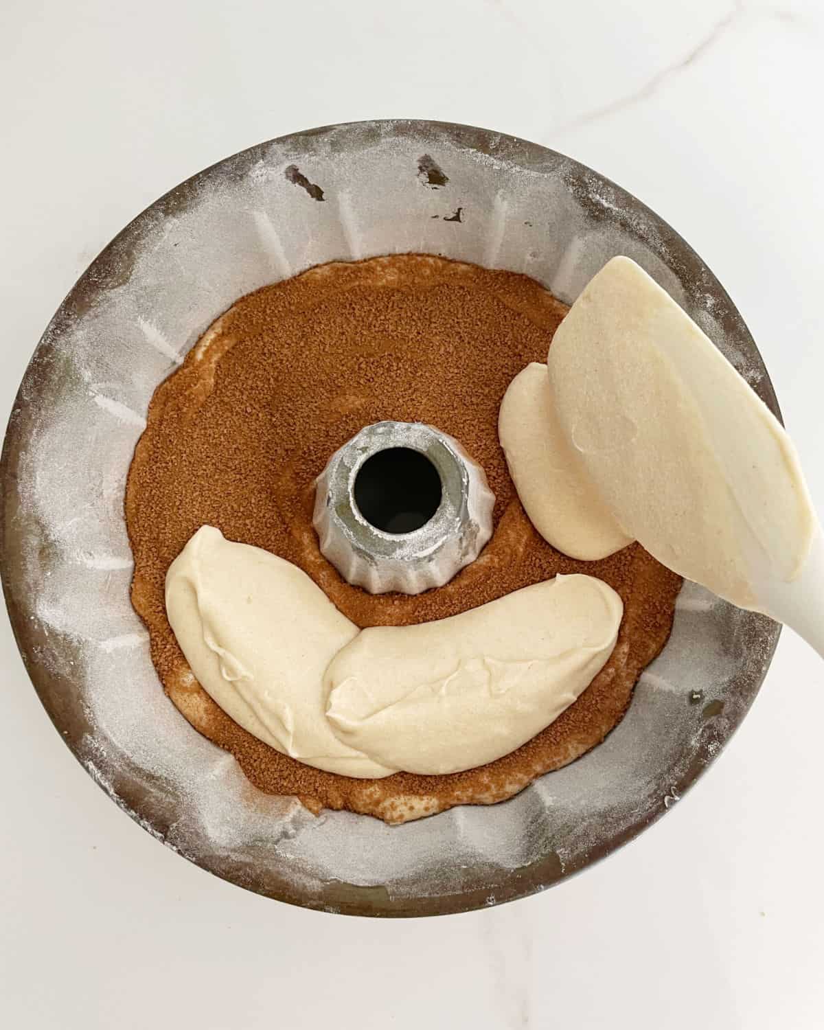 Layering cake batter and cinnamon sugar with a white spatula in a bundt pan on a white surface.