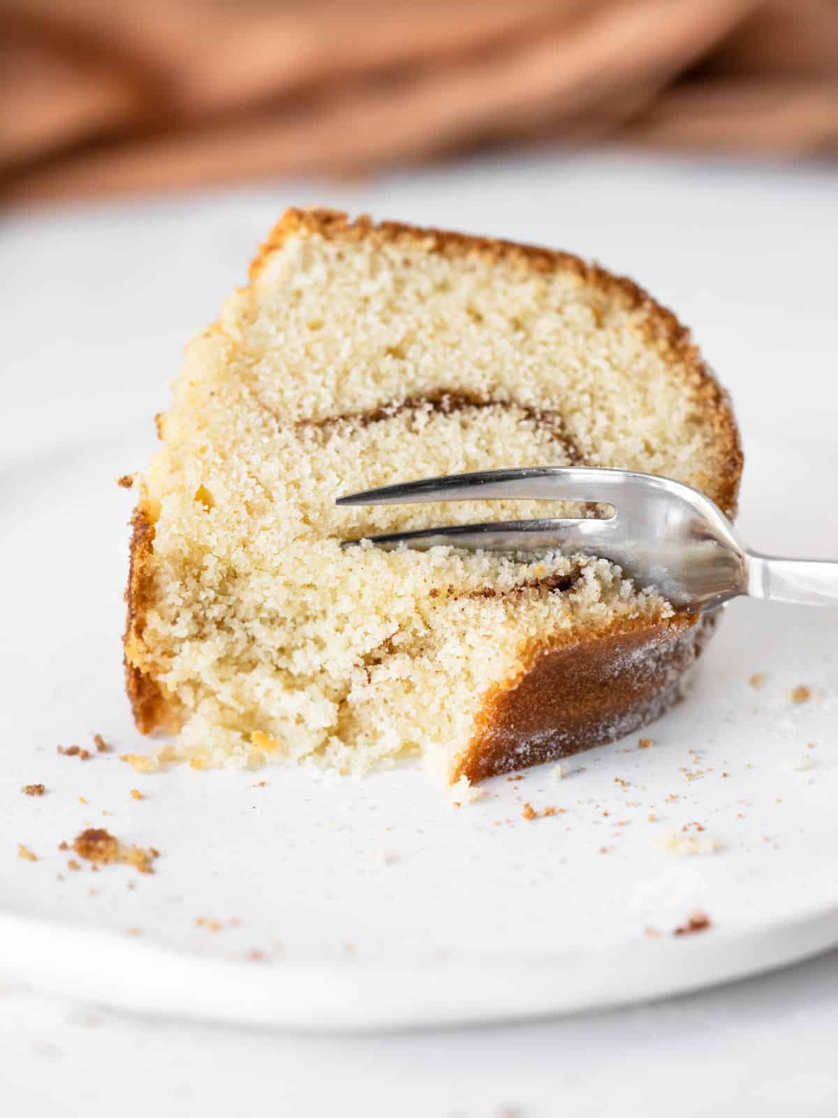 Forking a slice of cinnamon swirl bundt cake on a white plate. Brown cloth in the background.