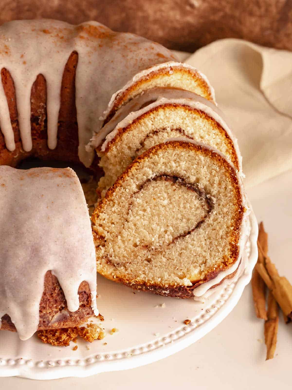 Several slices of glazed cinnamon swirl bundt cake on a white cake stand with rest of the cake.