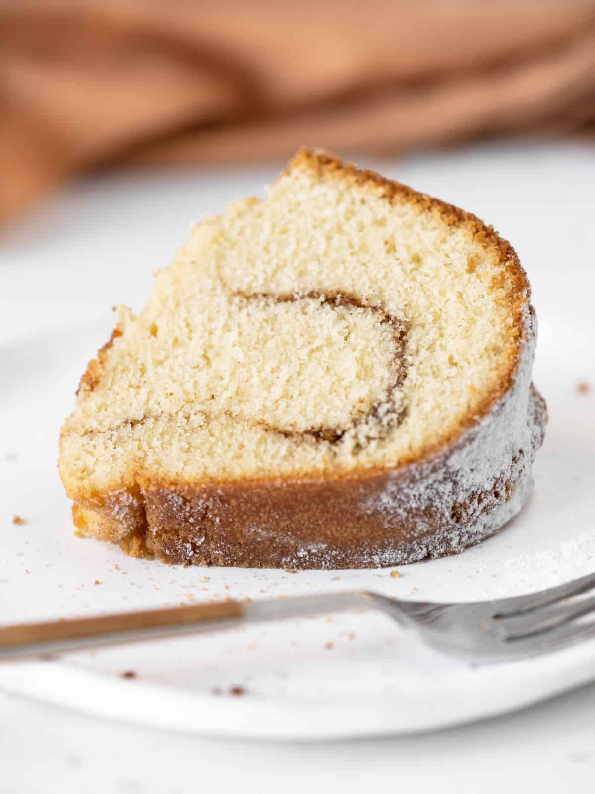 Single slice of cinnamon swirl bundt cake on a white plate with a wood and silver fork. Brown background.