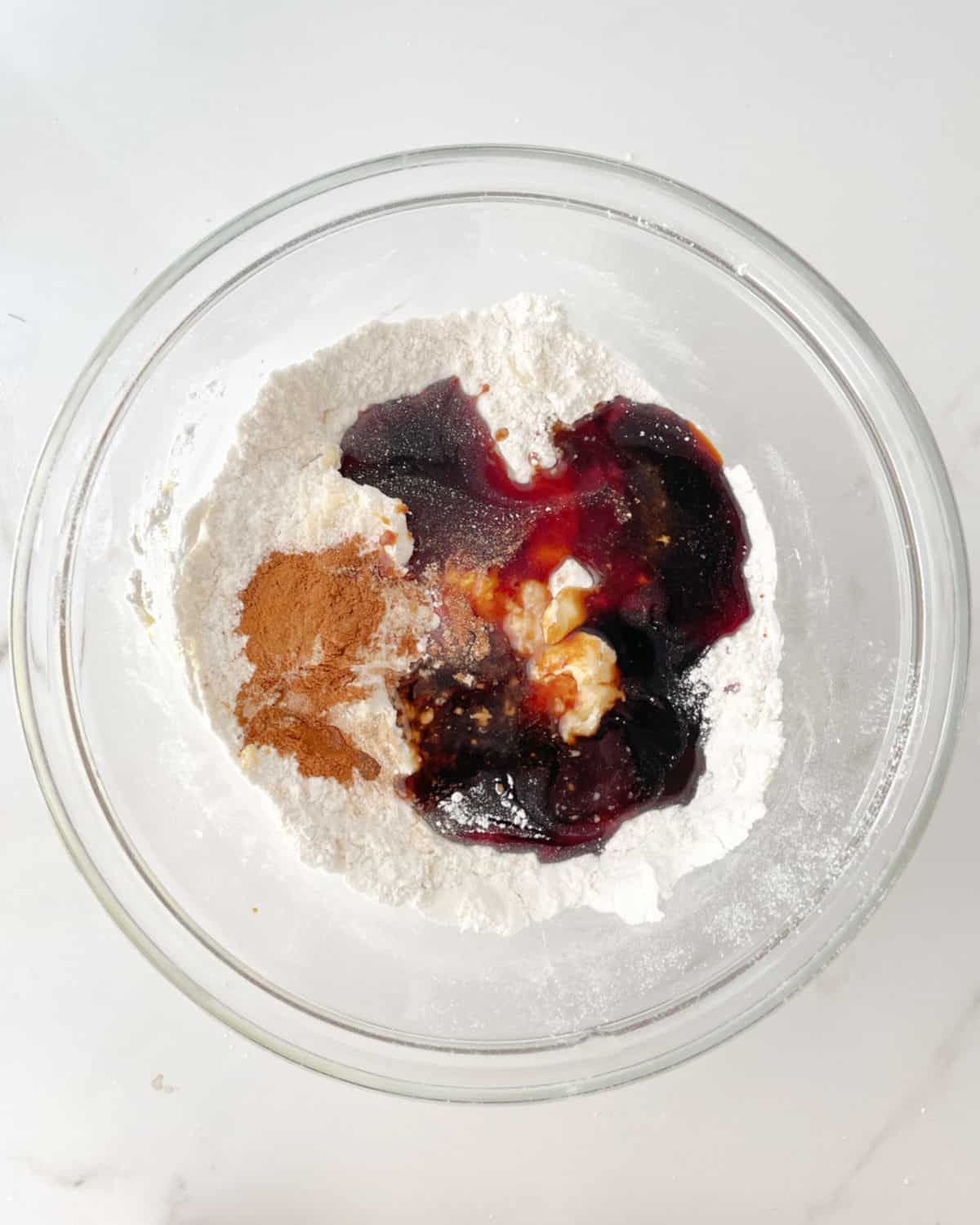 Glass bowl with flour, spices, butter, and molasses on a white marble surface. View from above.