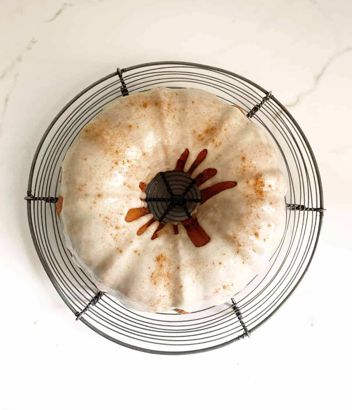 A wire rack with a glazed bundt cake on a white marble surface. View from above.
