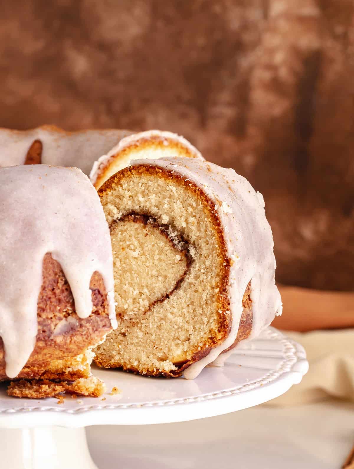 Glazed slice of cinnamon swirl bundt cake on a white cake stand with rest of cake. Brown background.