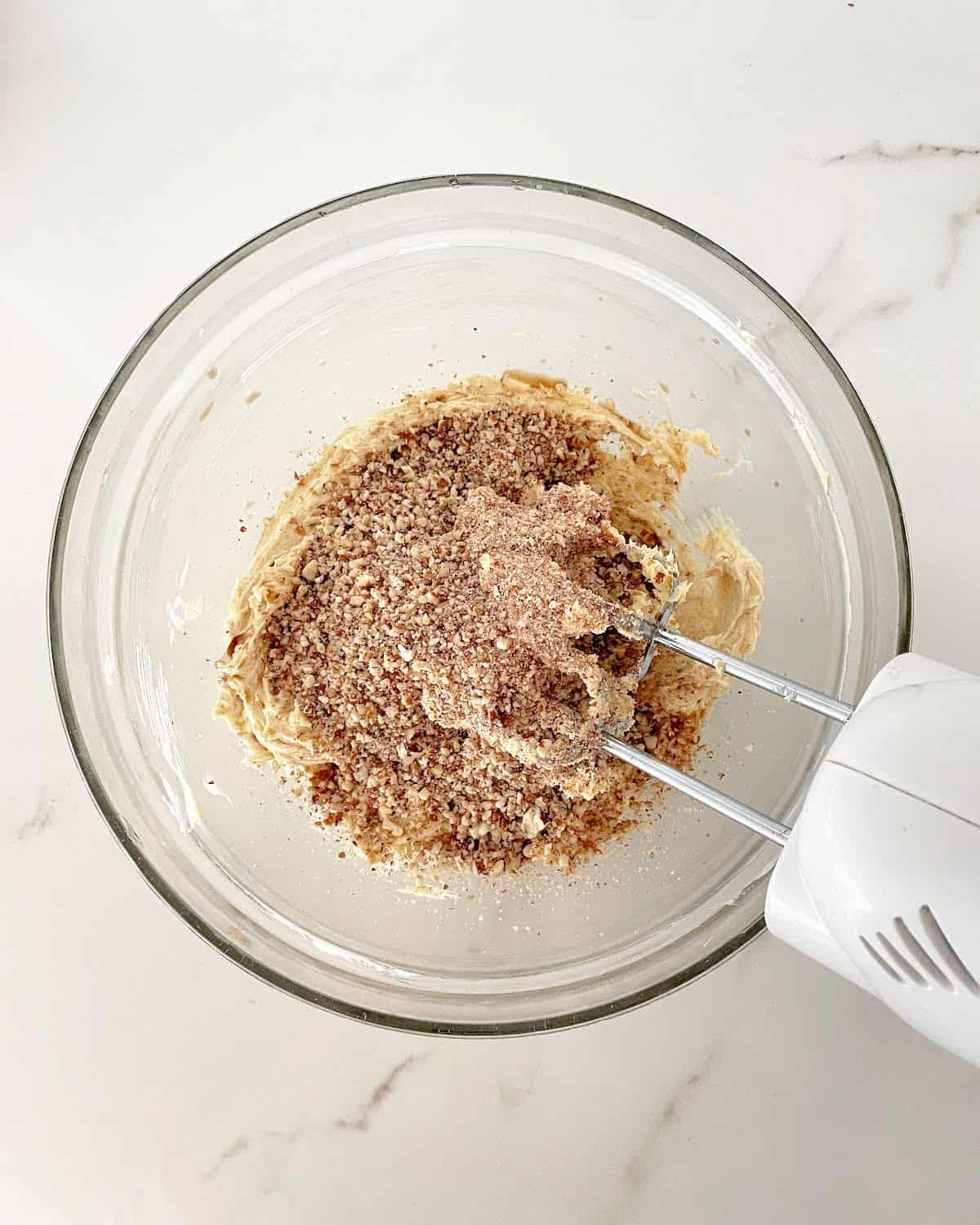 Ground pecans added to butter mixture in a glass bowl with an electric mixer. White marble surface.