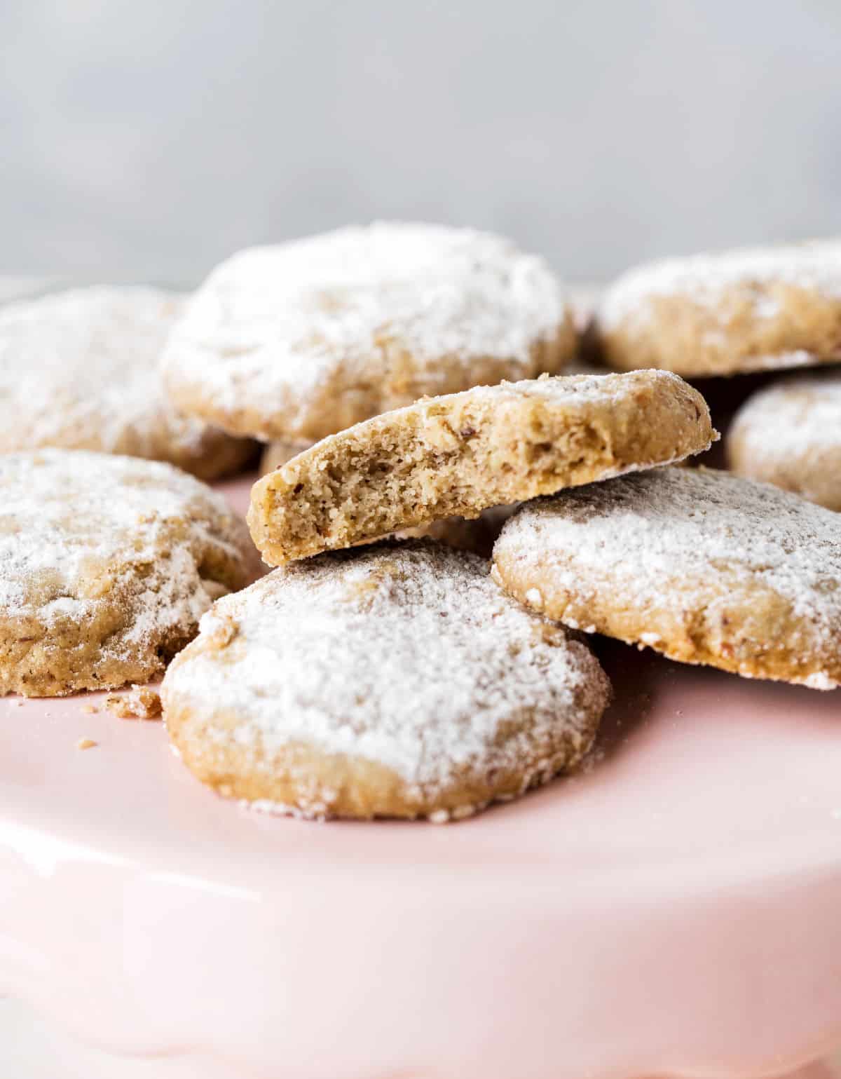 Pile of pecan cookies with powdered sugar, one is bitten, on a pink plate with a grey background,