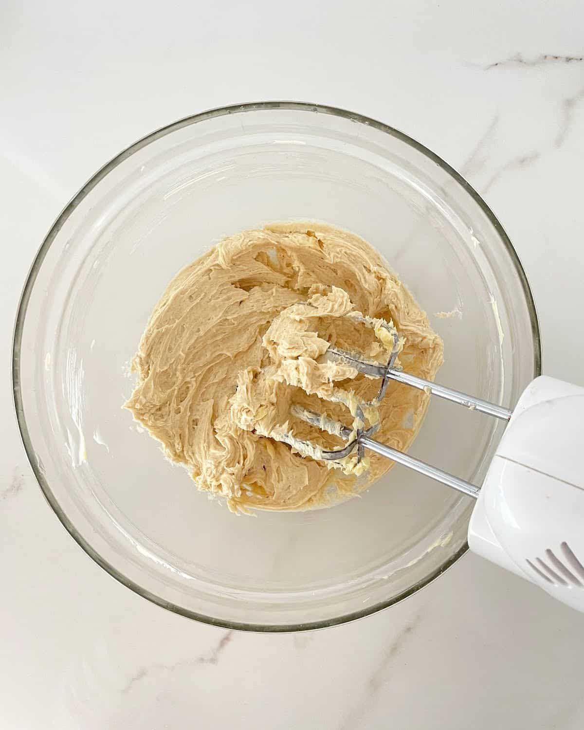 Beating brown sugar and butter in a glass bowl on a white marble surface.