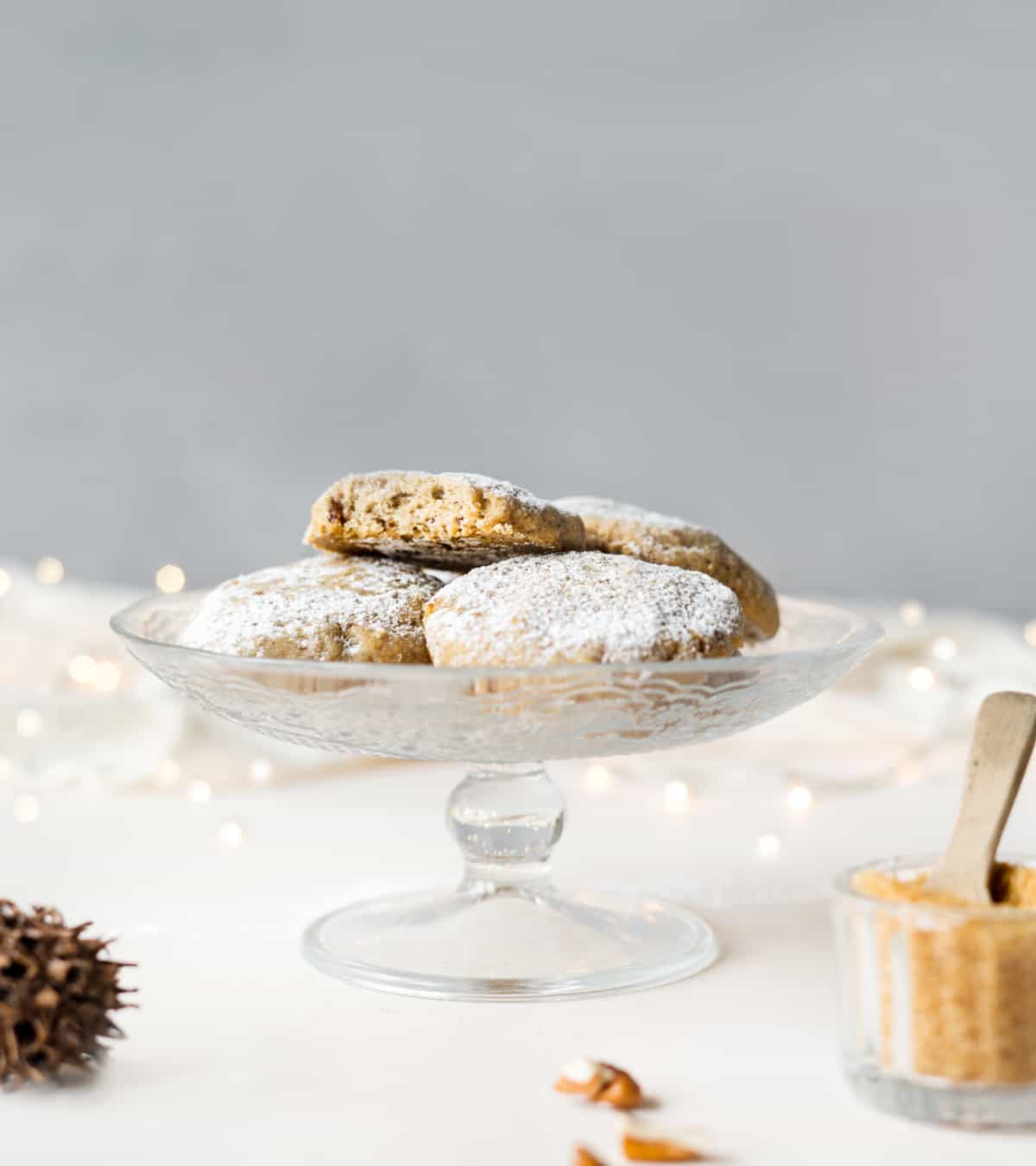 Glass cake stand with several pecan cookies with powdered sugar on a whitish surface with grey background.