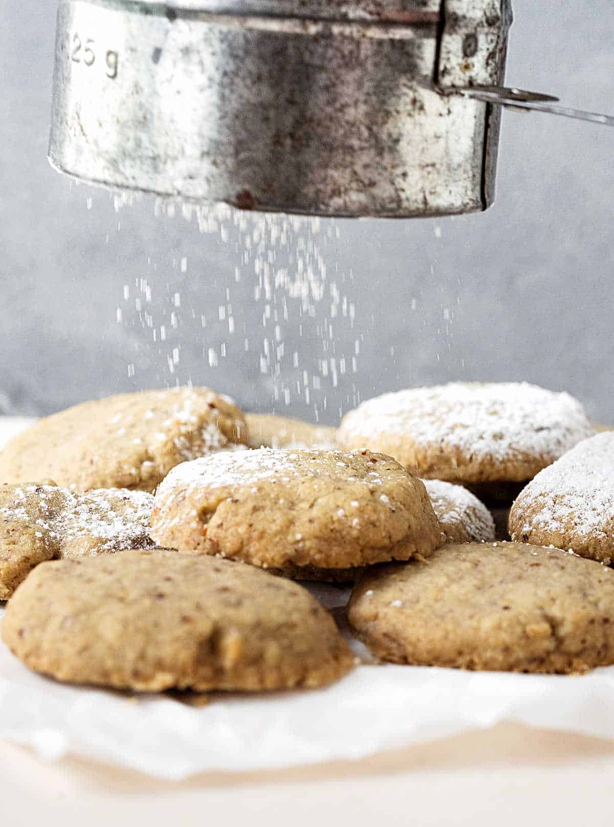 Powdered sugar being sifted over pecan cookies in a pile on a cream colored surface.