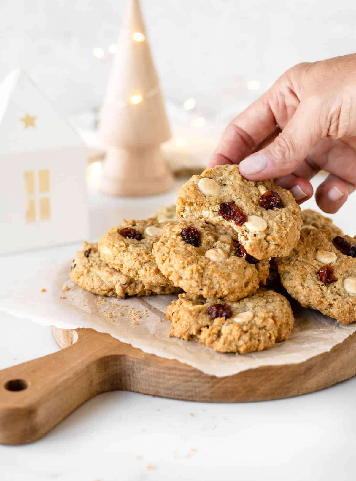 Wooden board with pile of cranberry white chocolate oatmeal cookies. A hand lifting a bitten one. White decorations with lights in the background. 