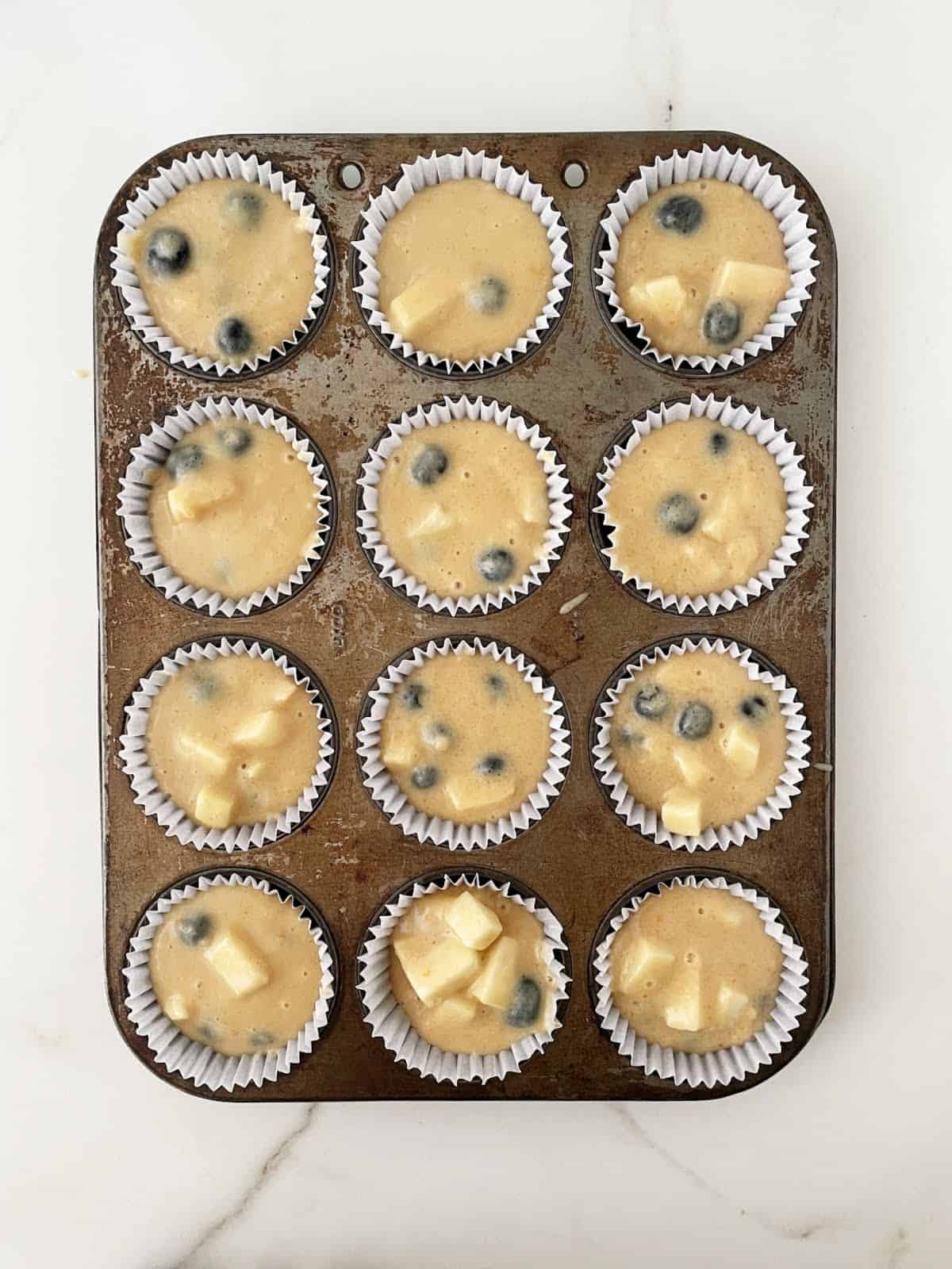 Blueberry apple muffins batter in paper cups in a metal muffin pan. White marble surface. Top view.