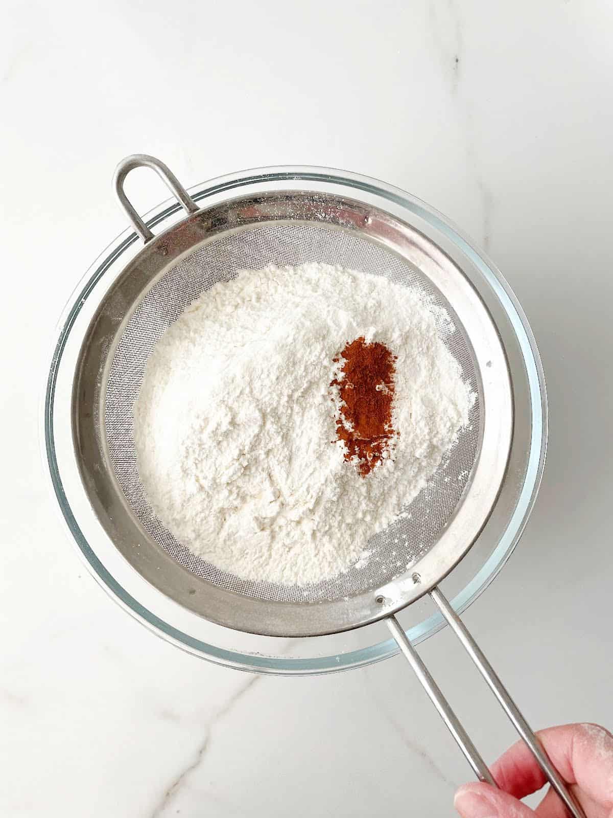 Sifting flour mixture and cinnamon over a glass bowl set on a white marble surface.